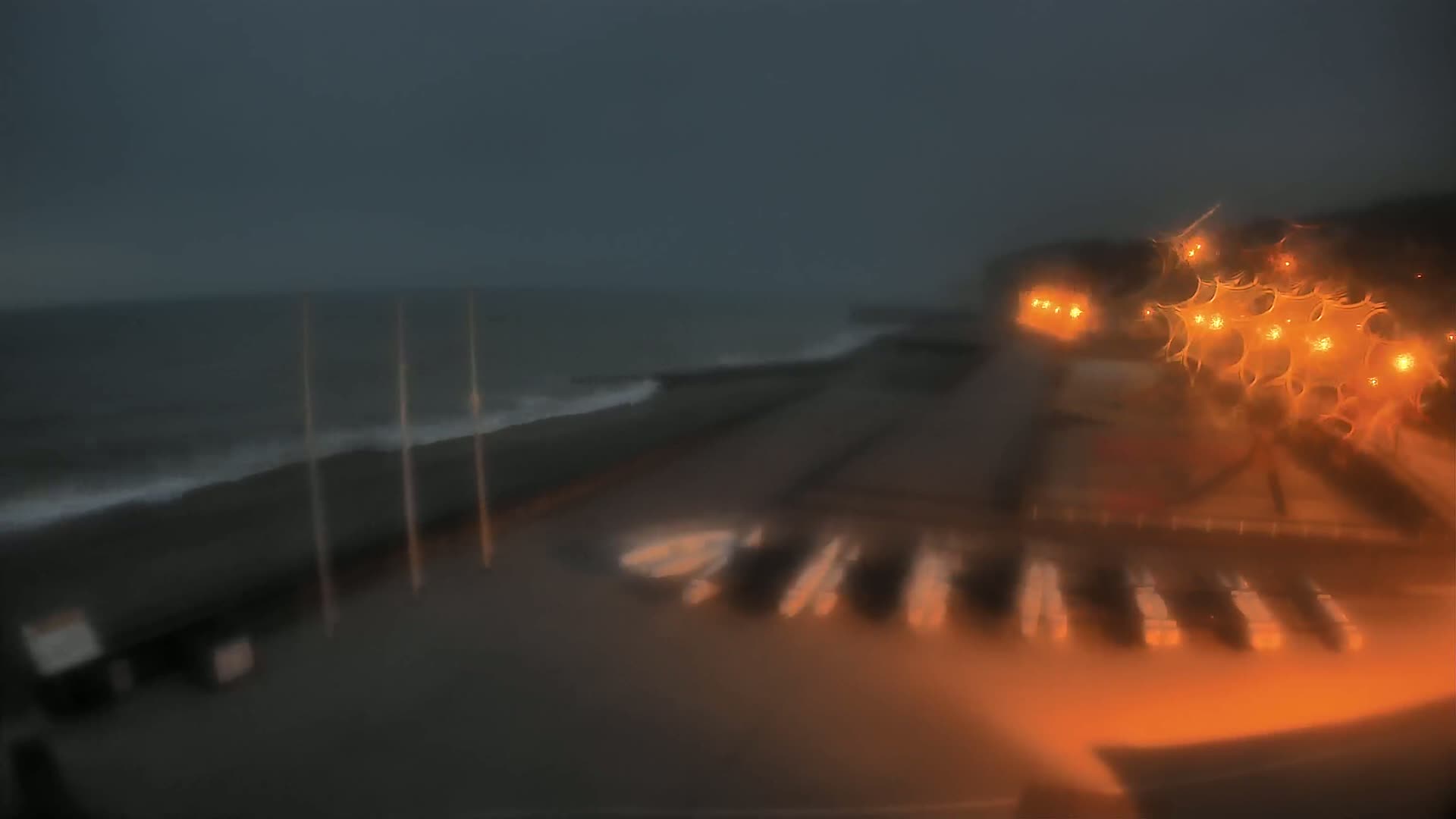 A blurry, nighttime view of a rain-soaked coastline shows a dark ocean with small waves, a shoreline with several boats, and illuminated buildings in the distance.