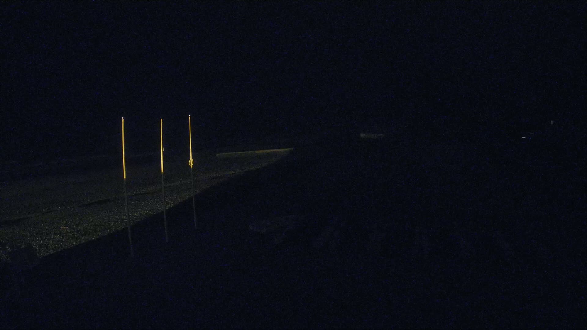 Three tall, thin, illuminated posts stand on a dark, gravelly surface at night.