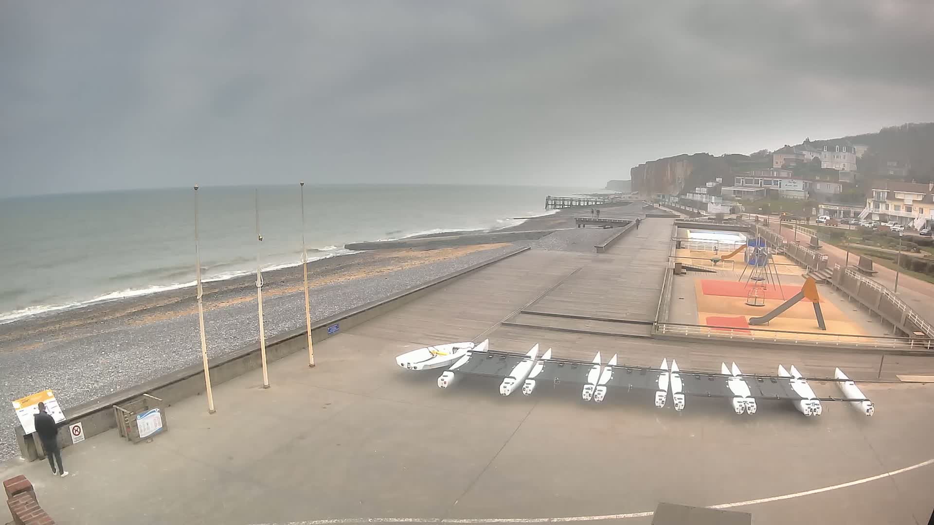 A mostly cloudy day shows a beach with a wooden boardwalk, several small sailboats, a playground, and a pier extending into a gray sea with small waves crashing on the shore.