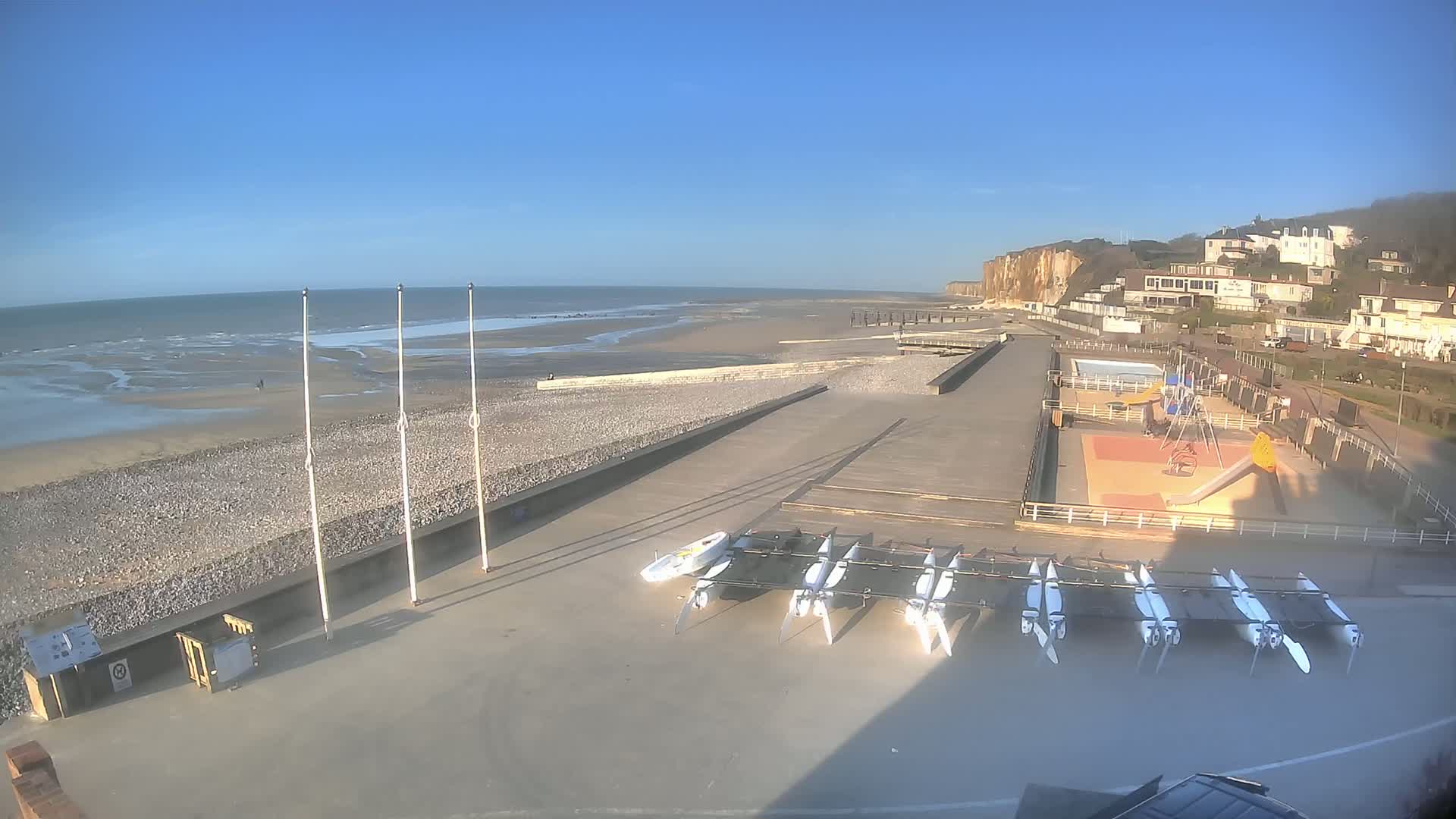 A beach with several small sailboats stored near a playground, a boardwalk, and white cliffs under a clear blue sky.