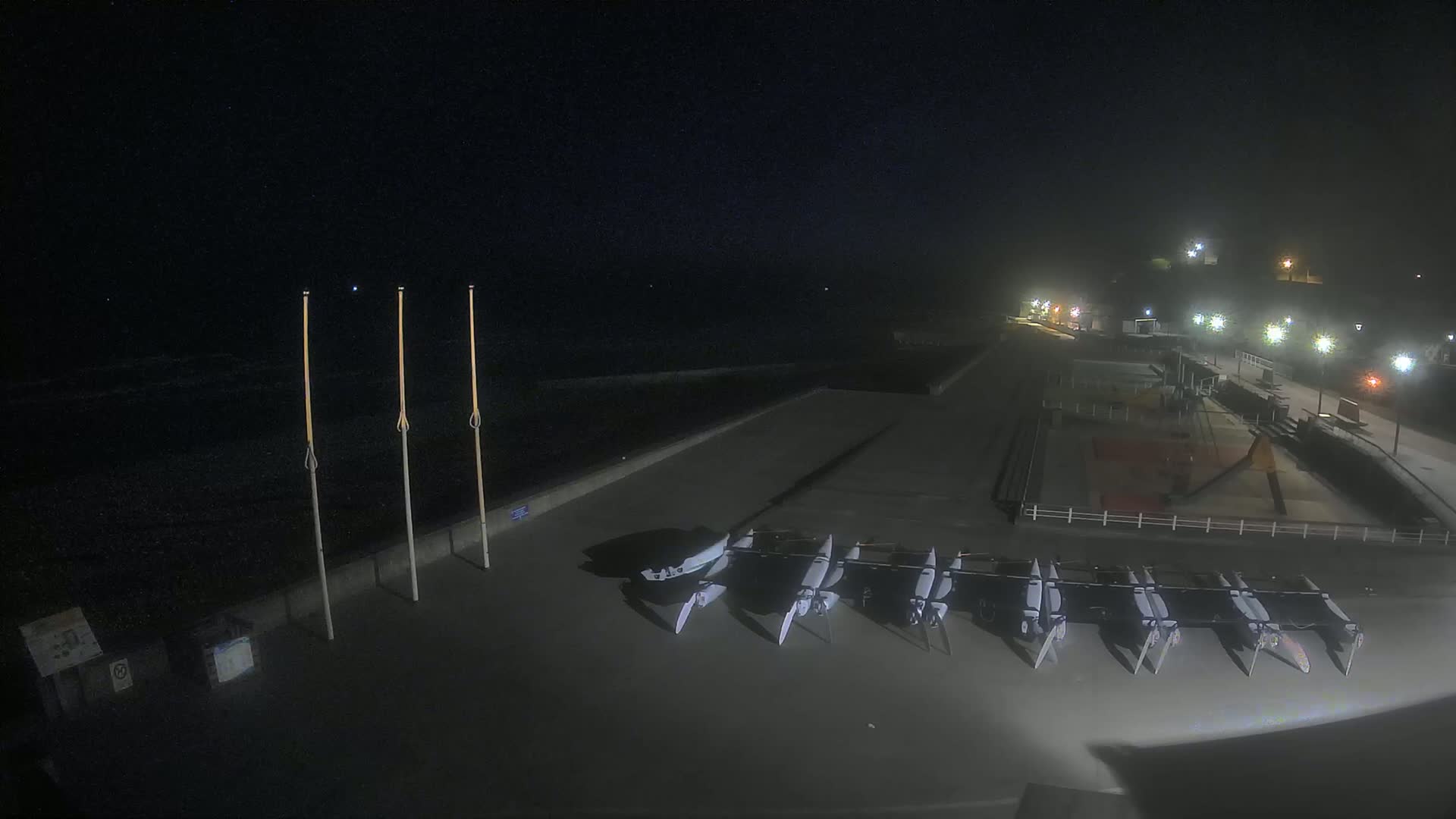 At night, several small sailboats are stored on a paved waterfront area next to a dark, wavy ocean under a hazy sky.