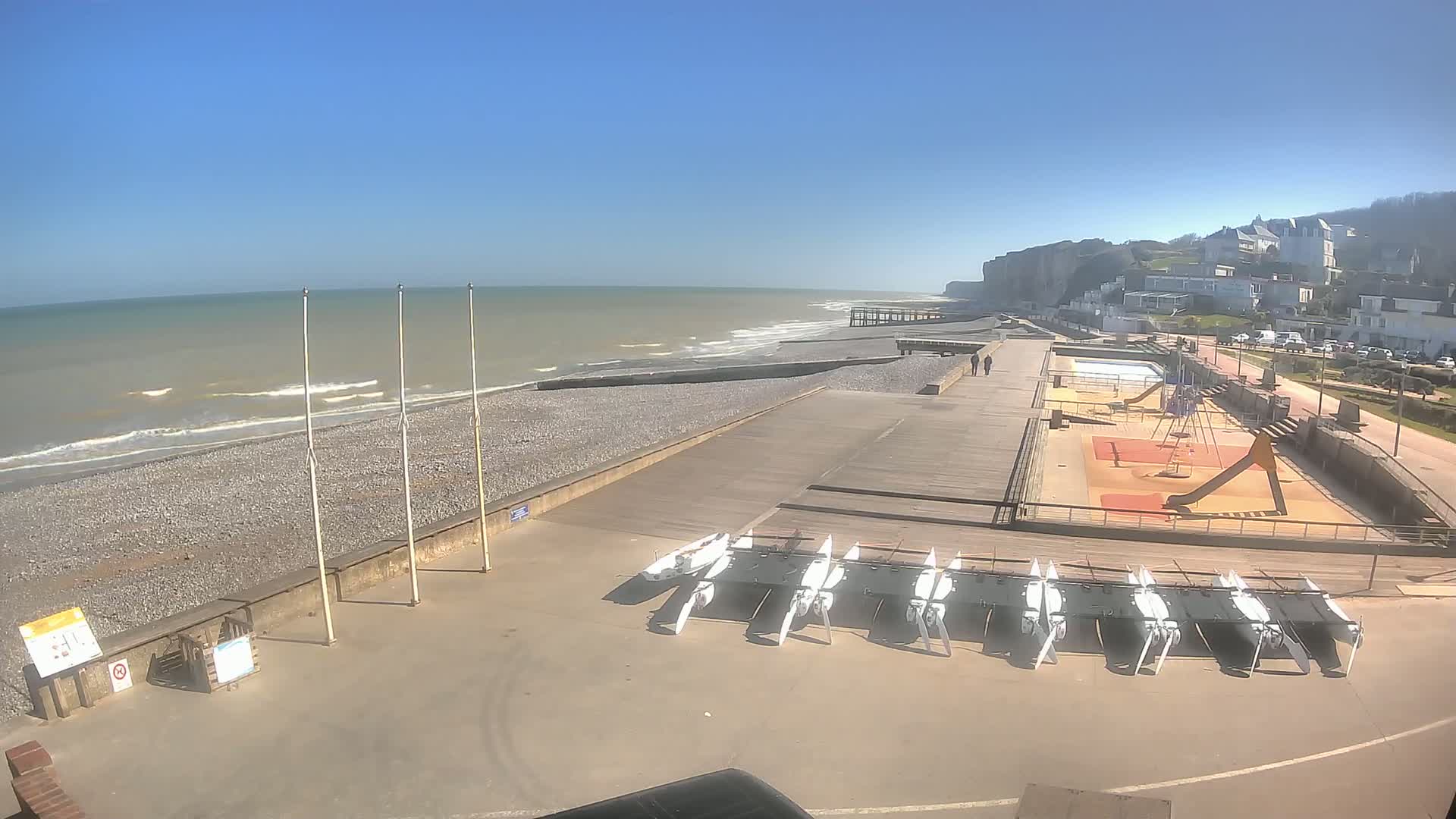 A pebble beach with a boardwalk, playground, and several small sailboats is visible under sunny skies, next to white cliffs and a town.