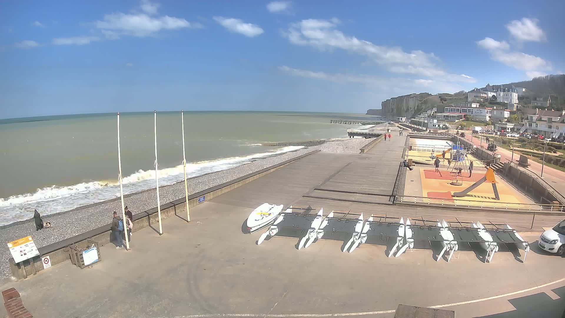 A beach with a pebble shoreline, small waves, a wooden boardwalk, a playground, several small sailboats, and white cliffs in the background under a mostly sunny sky.