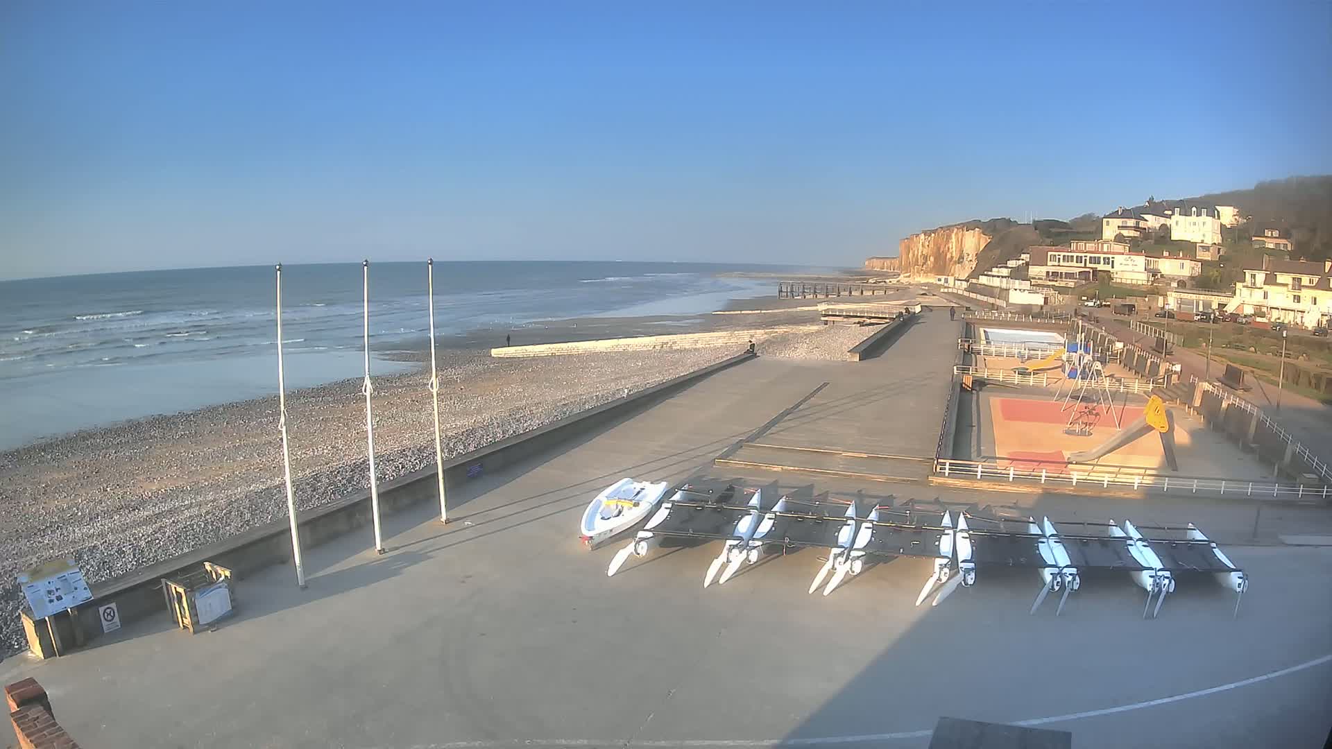 A mostly sunny day shows a pebbly beach with calm waves, several small sailboats parked on a paved area, a playground, and white cliffs in the background.