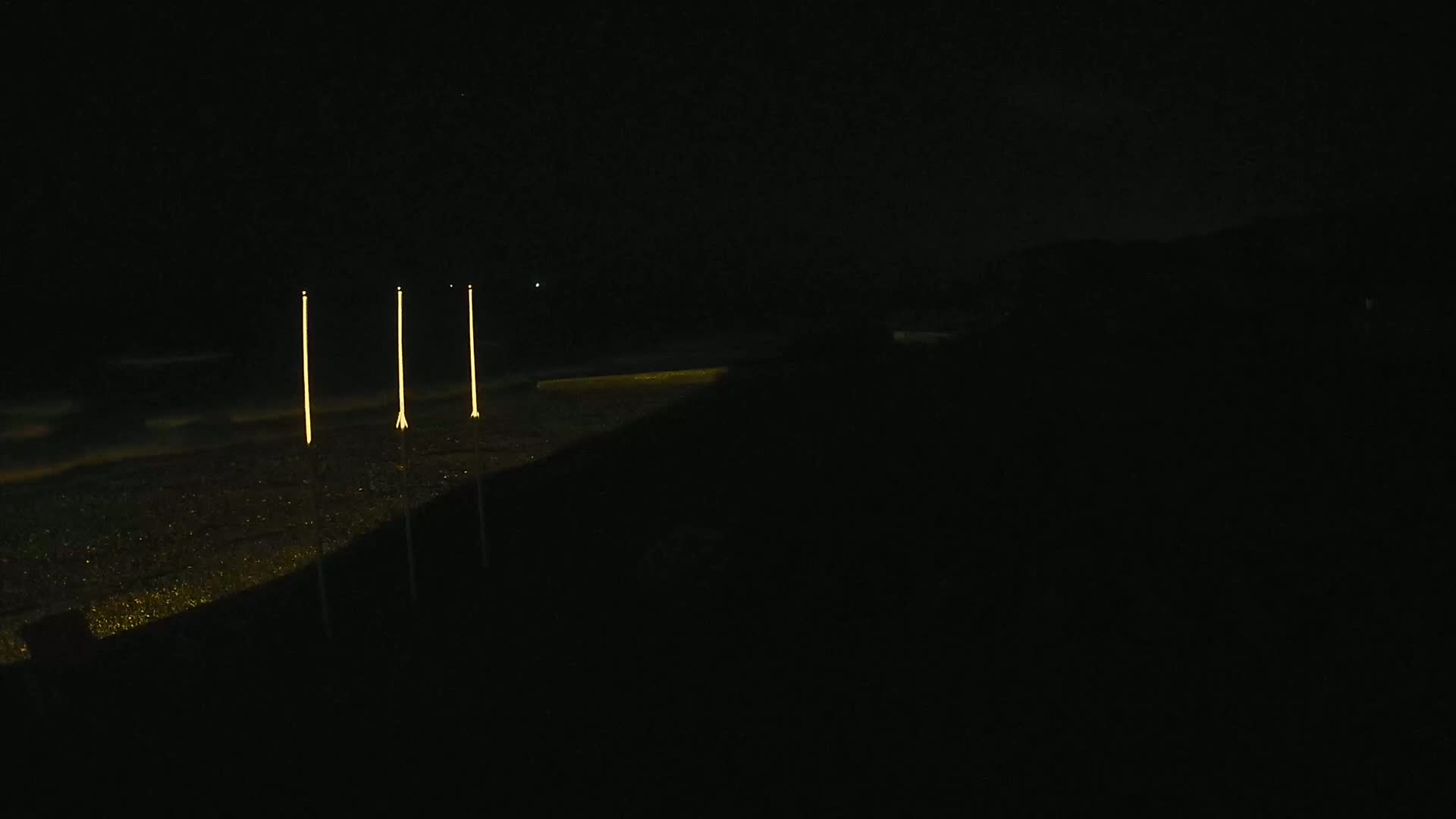 Three tall, slender lights illuminate a dark, seemingly sandy shoreline at night.