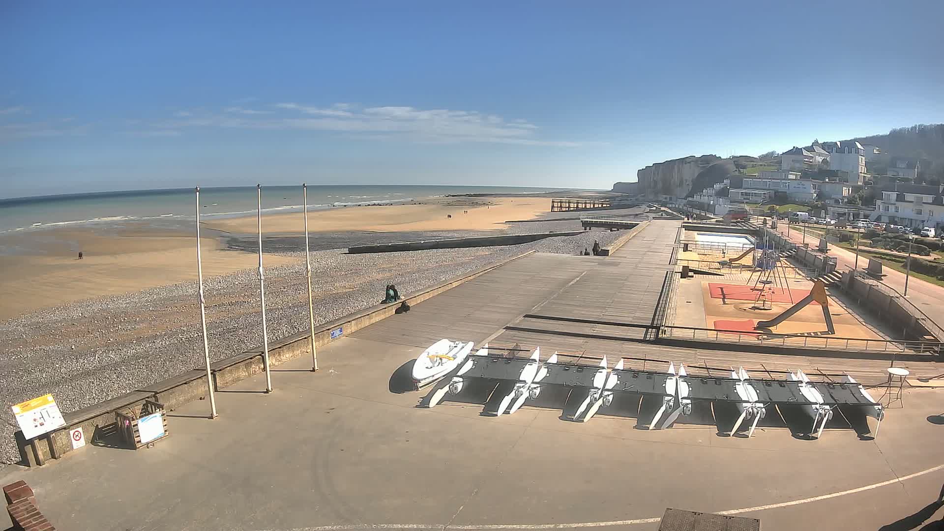 A sunny day at a beach with a wooden boardwalk, a playground, and several small sailboats stored on the ground.