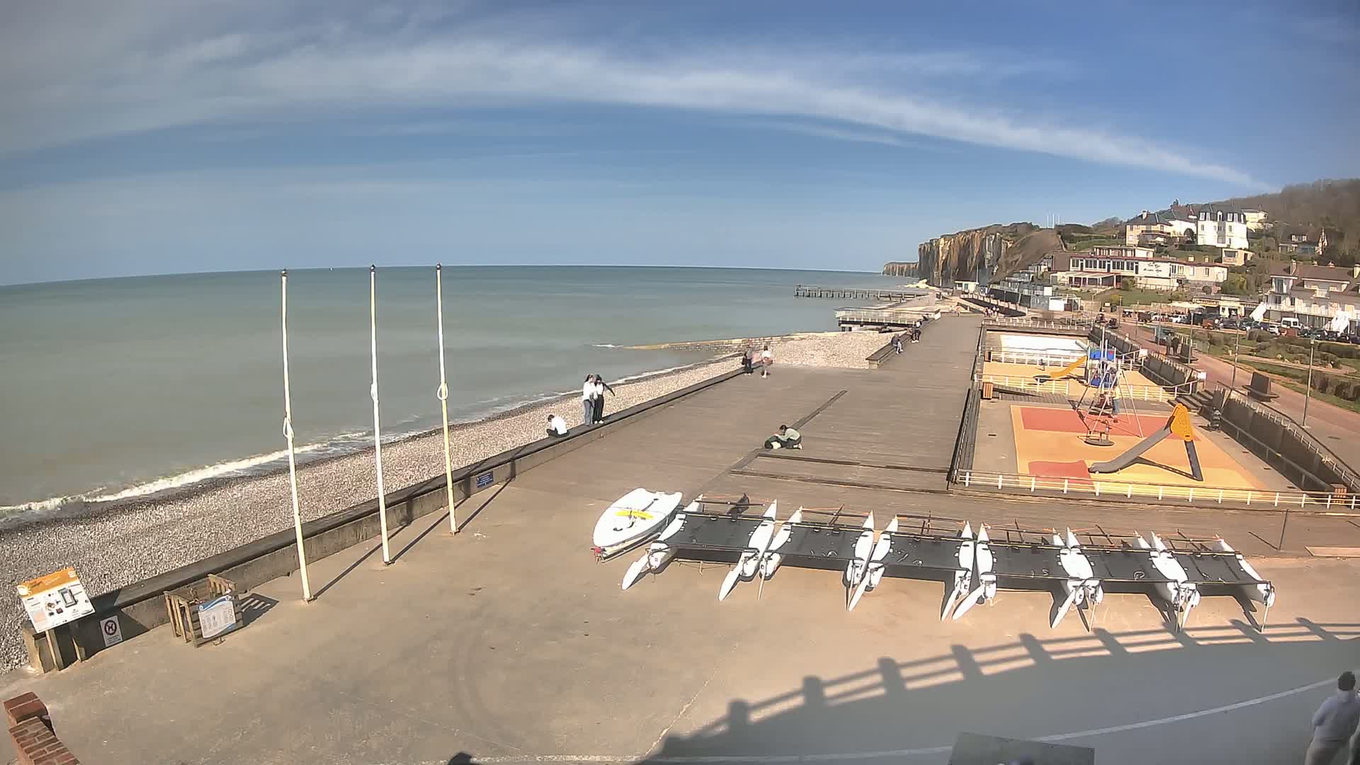 A beach with a boardwalk, playground, and several small sailboats is visible under a mostly sunny sky; white-capped waves lap the shore.