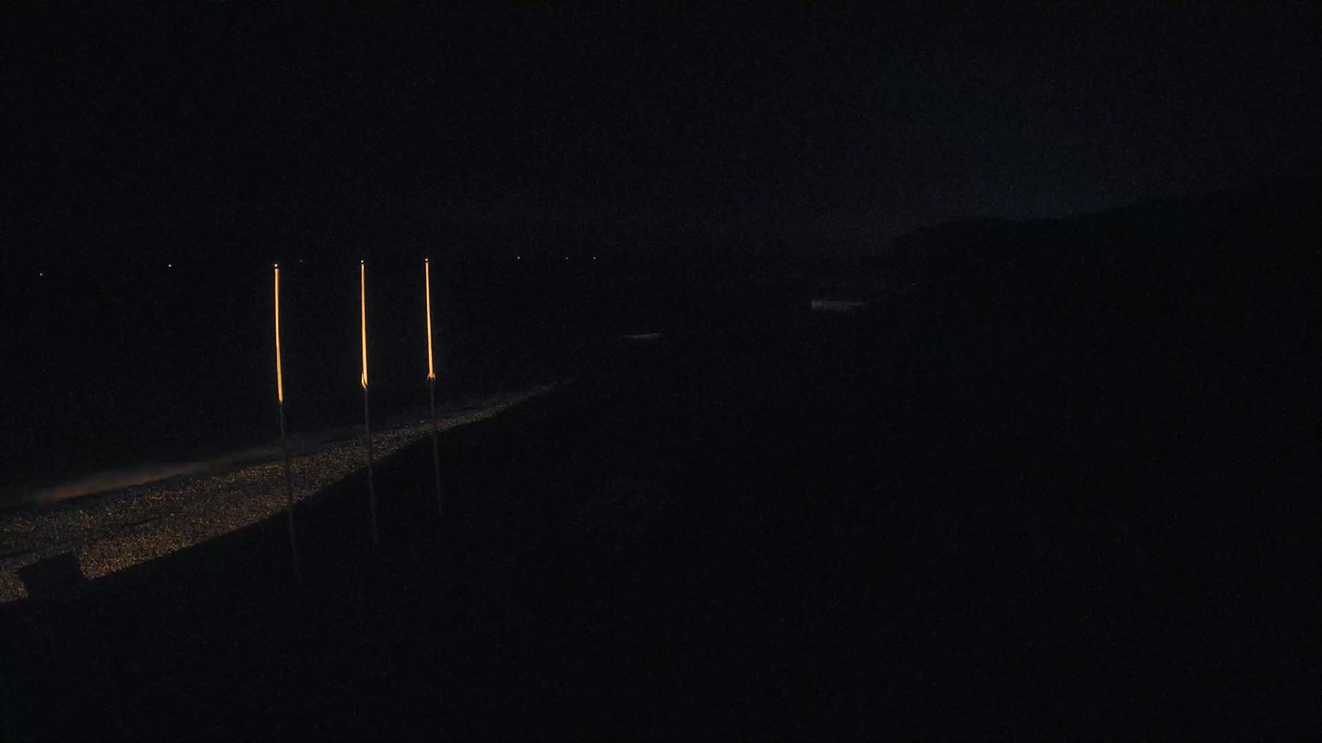Three tall, slender lights illuminate a dark, gravelly area at night.