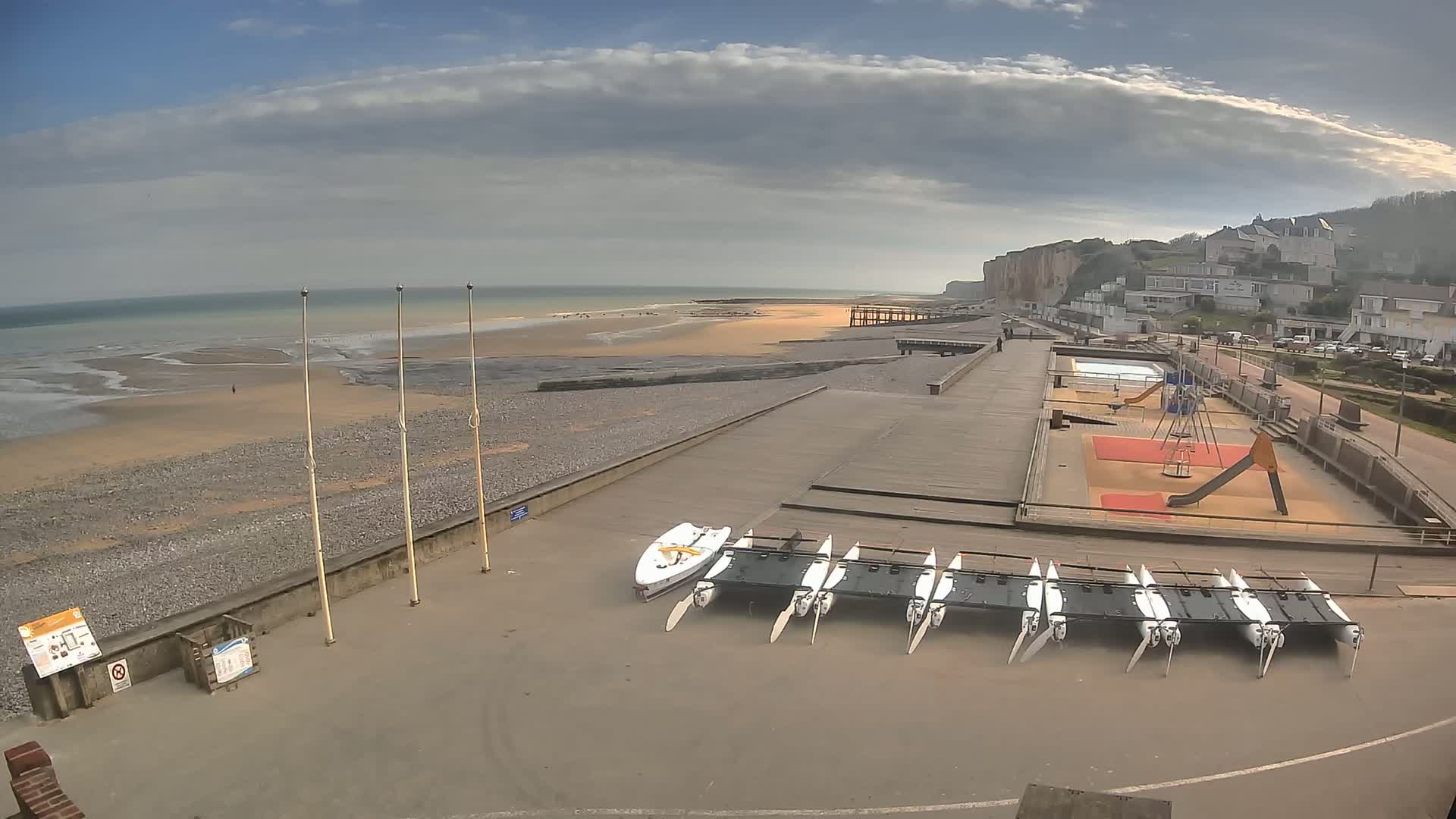 A mostly sunny day at a beach with a pebble shore shows several small sailboats stored near a playground, a boardwalk, and white cliffs in the background.