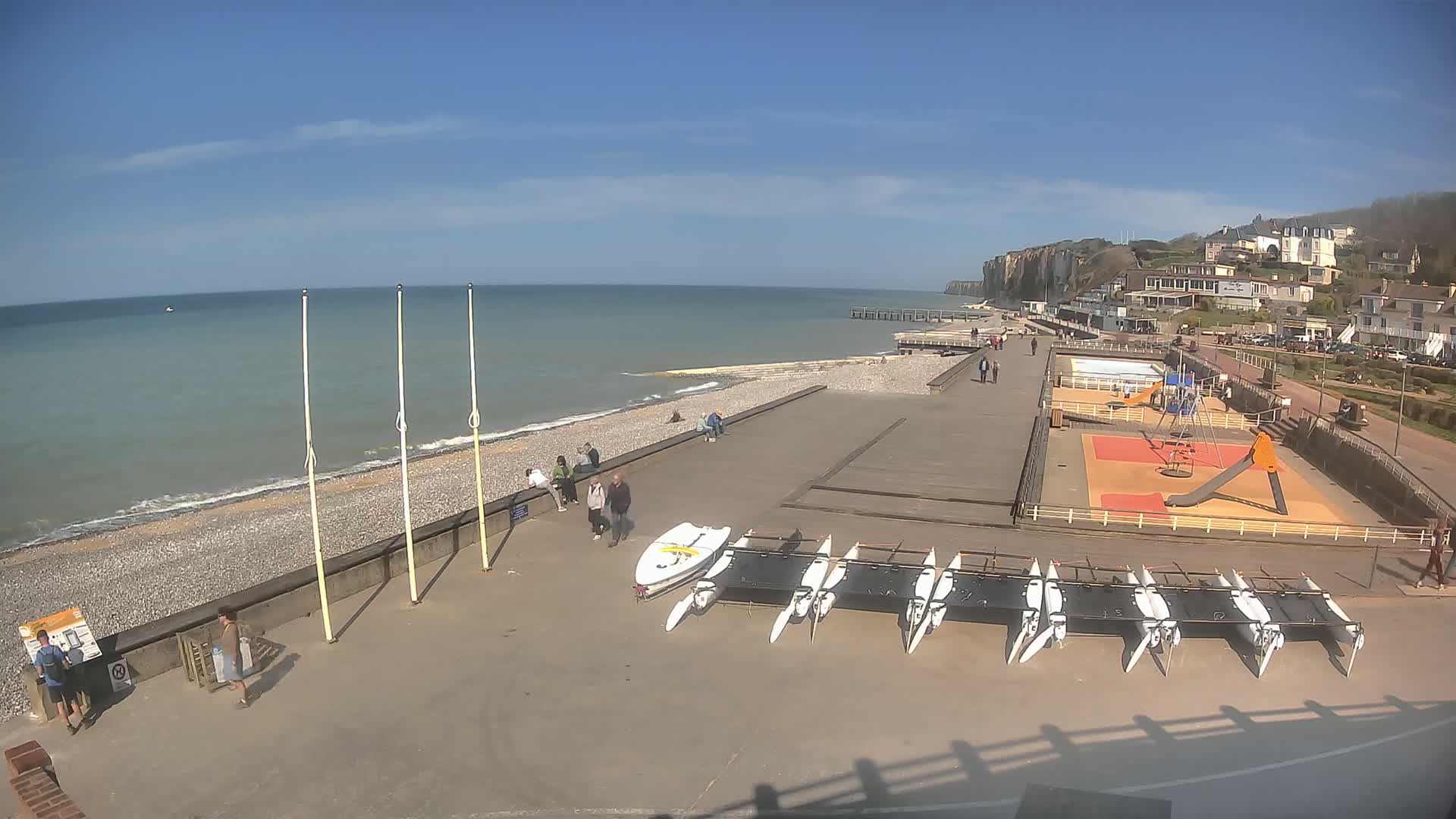 A sunny day at a seaside town reveals a pebble beach, a boardwalk with several people walking, a playground, and a row of catamarans, all set against a backdrop of white cliffs and the calm blue sea.