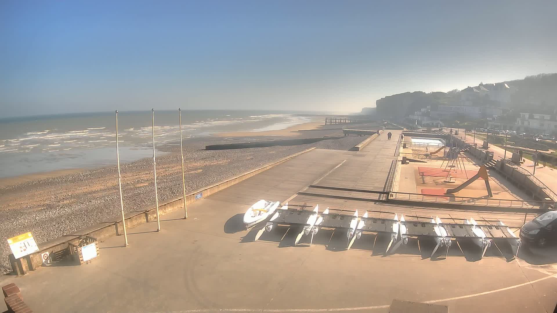 A beach with calm waves under a clear blue sky features a paved boardwalk with several small sailboats and a playground next to a cliffside town.