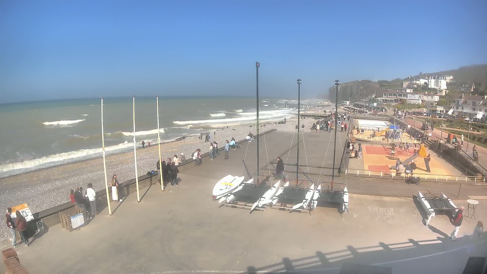 A beach scene on a sunny day with moderately wavy ocean water, showing people relaxing and playing on the beach and a playground, and several catamarans stored on the shore.