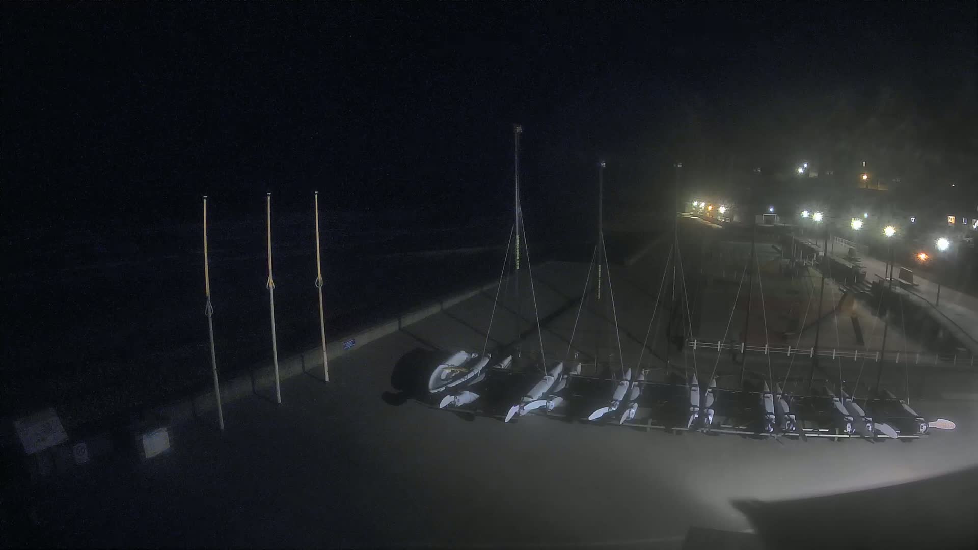 At night, several small sailboats are stored on a dark paved area near a shoreline under dim lighting.