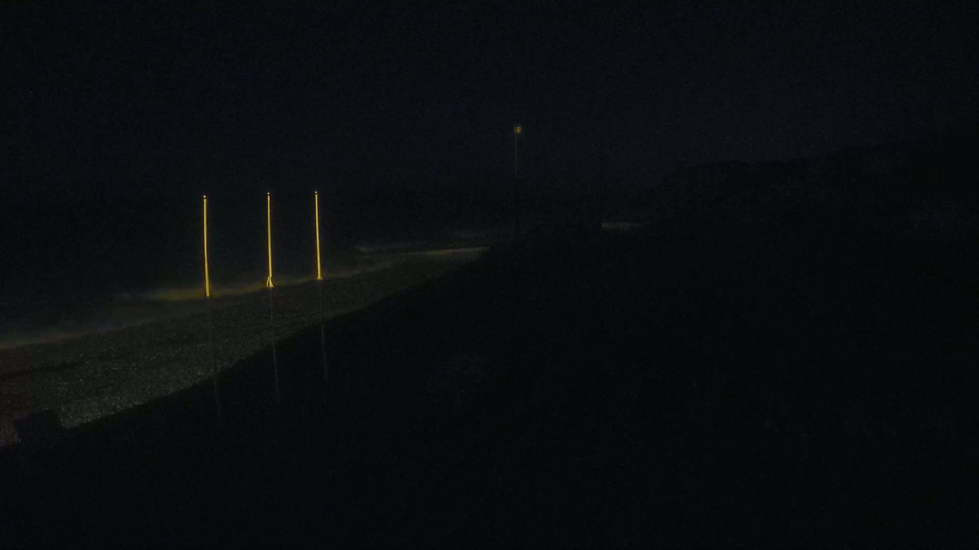 Three tall, thin lights illuminate a dark beach at night.