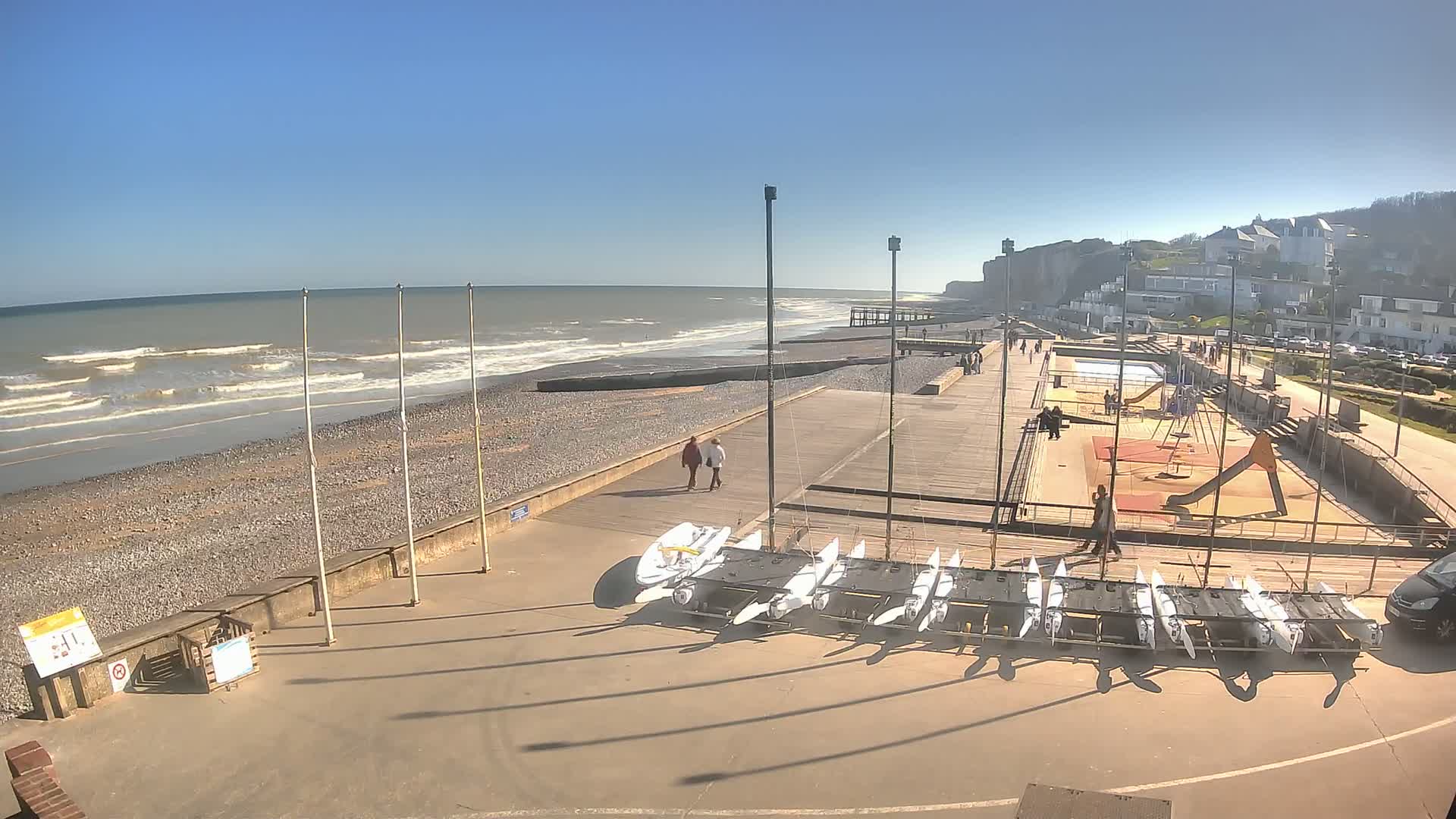 A seaside promenade on a sunny day with small waves, showing a pebble beach, several sailboats on trailers, a playground, and a few people walking along the boardwalk.