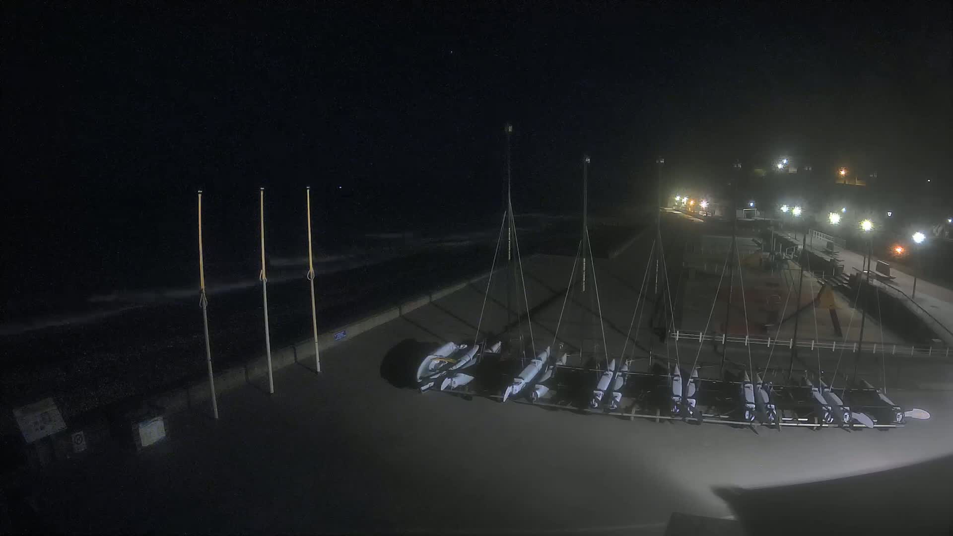 Several small sailboats are stored on a dark beach at night under hazy conditions, with lights illuminating a nearby walkway.