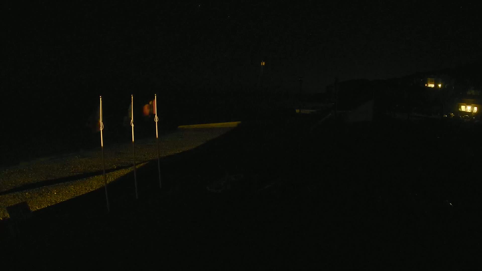 Three illuminated flags stand on a dark, pebble-covered surface at night, with dimly lit buildings visible in the background.