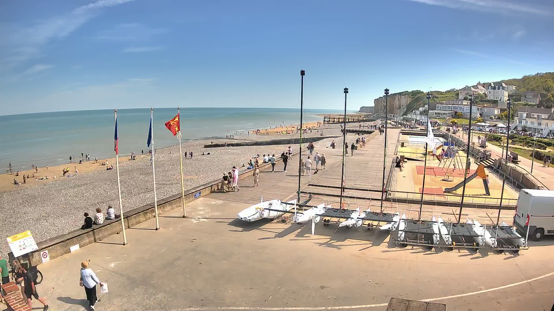 A sunny day at a pebble beach shows people relaxing and playing on the sand and a boardwalk, with several small sailboats parked nearby and a playground in the background.