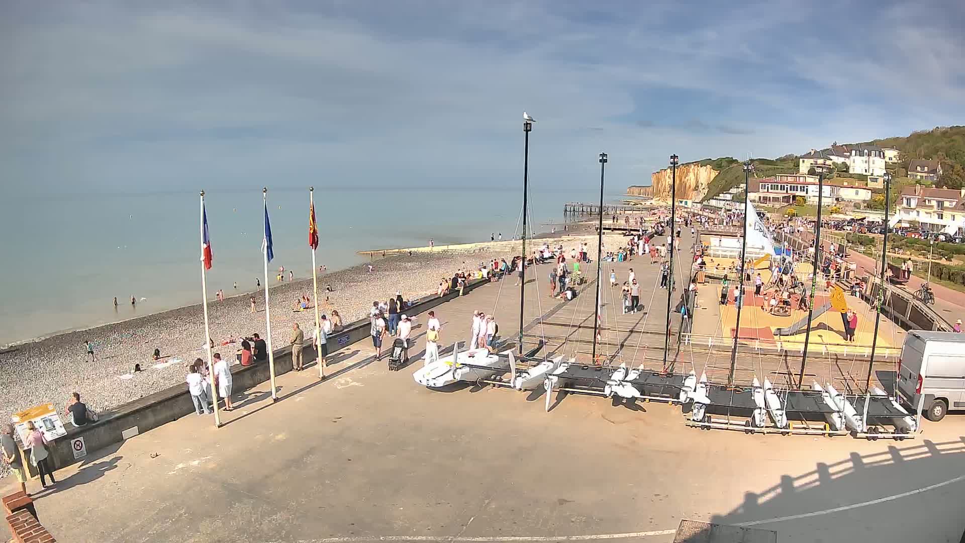 A pebble beach crowded with people on a sunny day, with sailboats and a playground next to a boardwalk leading to white cliffs.