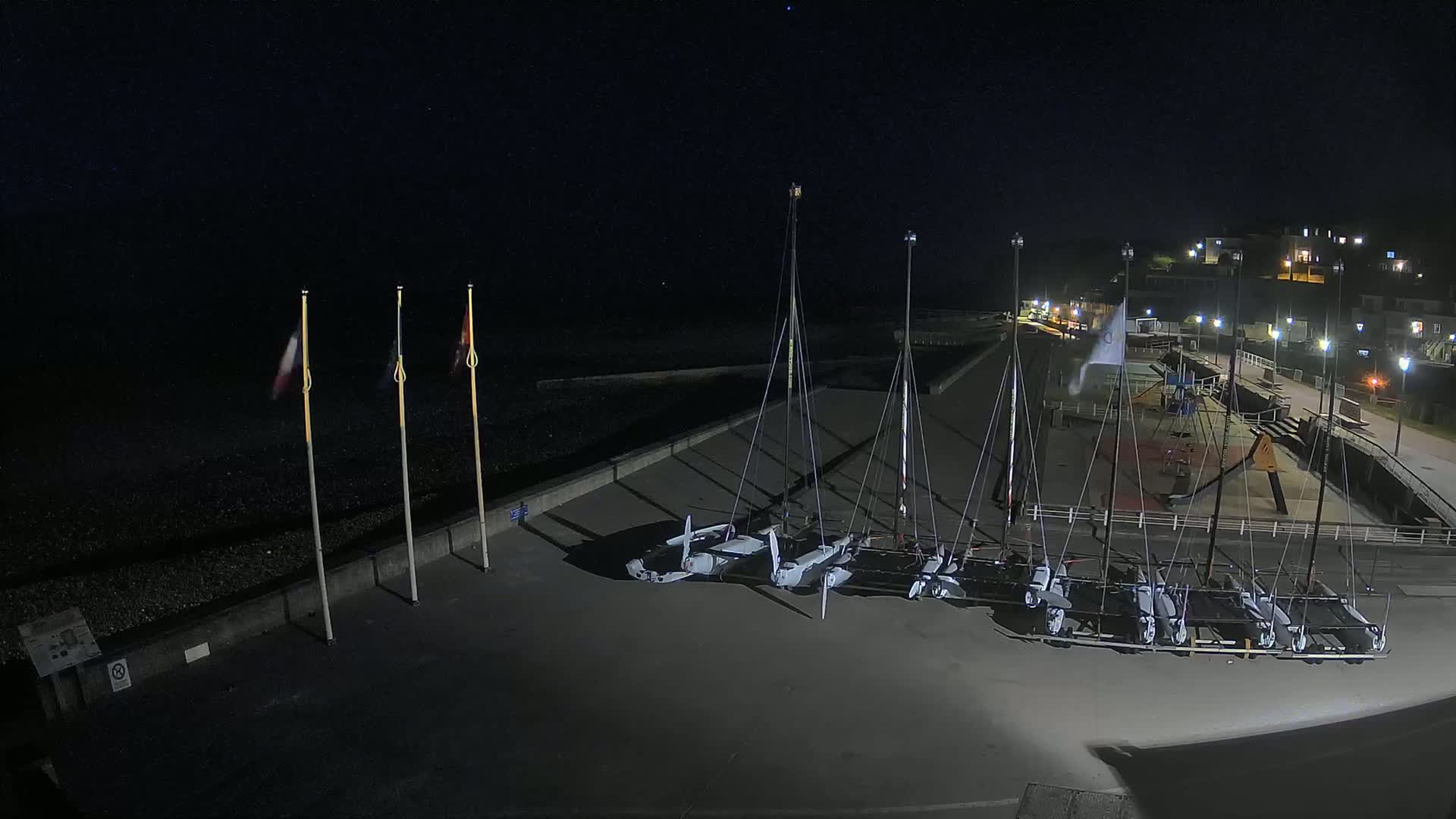 At night, several sailboats are docked on a paved area next to a pebble beach, under a clear, dark sky.