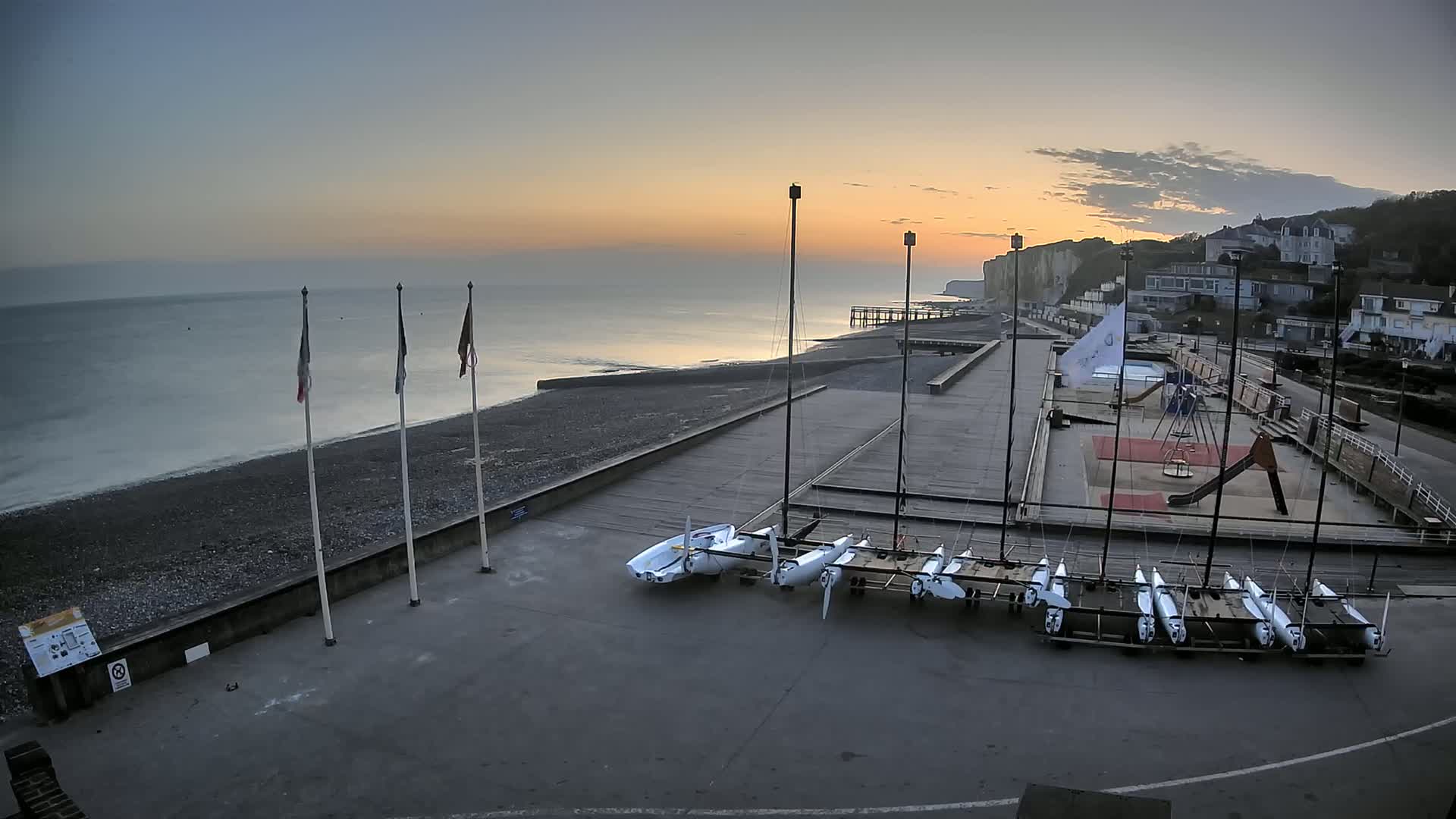 Several sailboats are stored on trailers next to a calm ocean at sunrise, near a playground and residential buildings on a cliff.