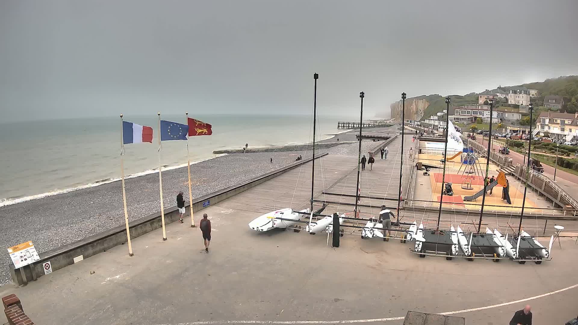 A rocky beach with several sailboats parked on a paved area, alongside a boardwalk, under an overcast sky;  people are visible walking along the shore and near the boats.