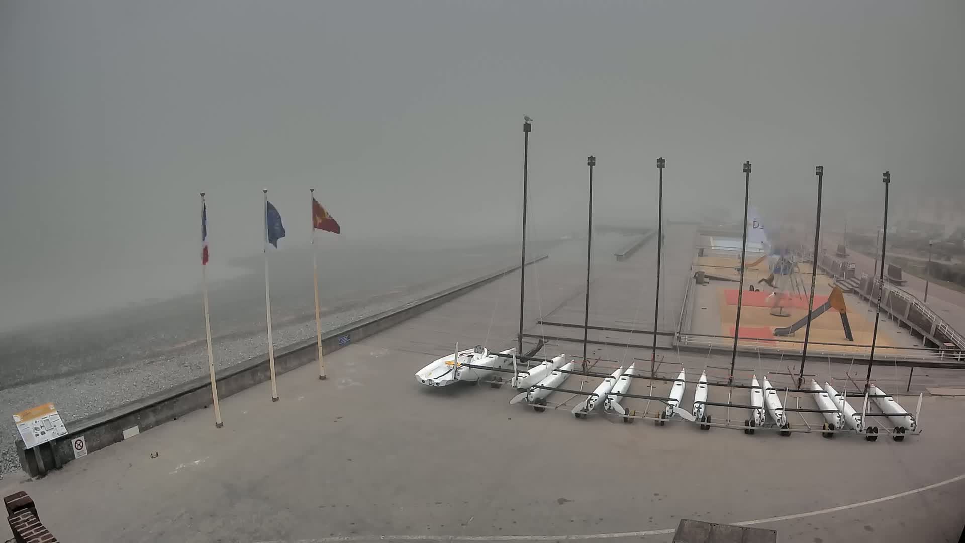 Several small sailboats are stored on a concrete area next to a rocky beach in foggy conditions.
