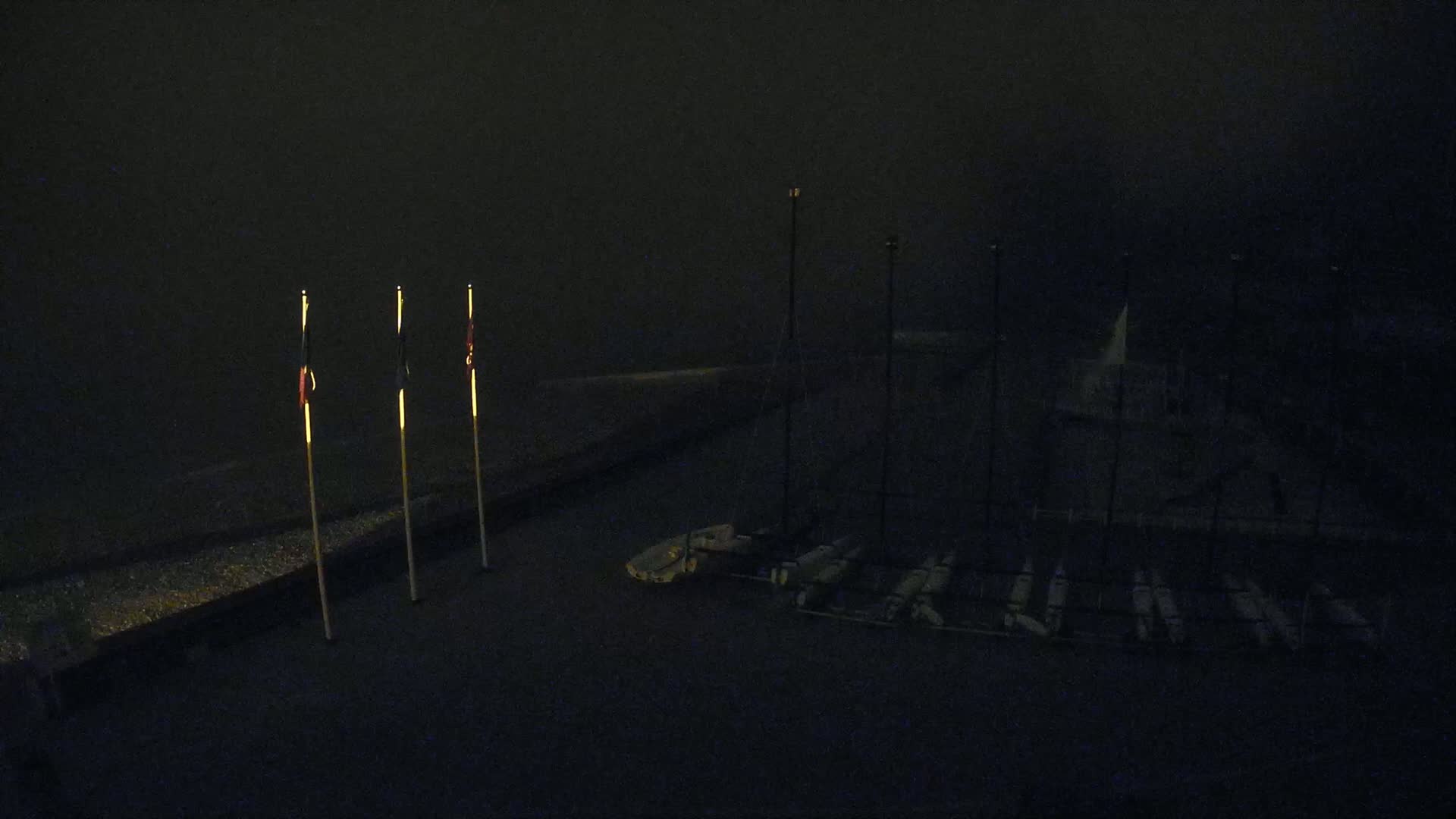 Three small flags are illuminated on poles at night, next to several small boats stored on racks in low-light conditions.
