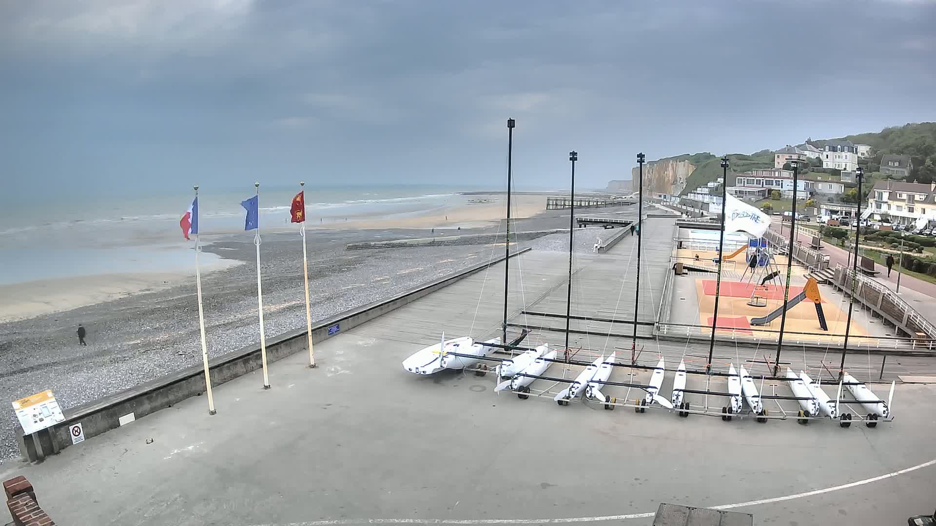 Several small sailboats are parked on a paved area next to a beach with a gray, overcast sky, and whitecaps on the ocean.