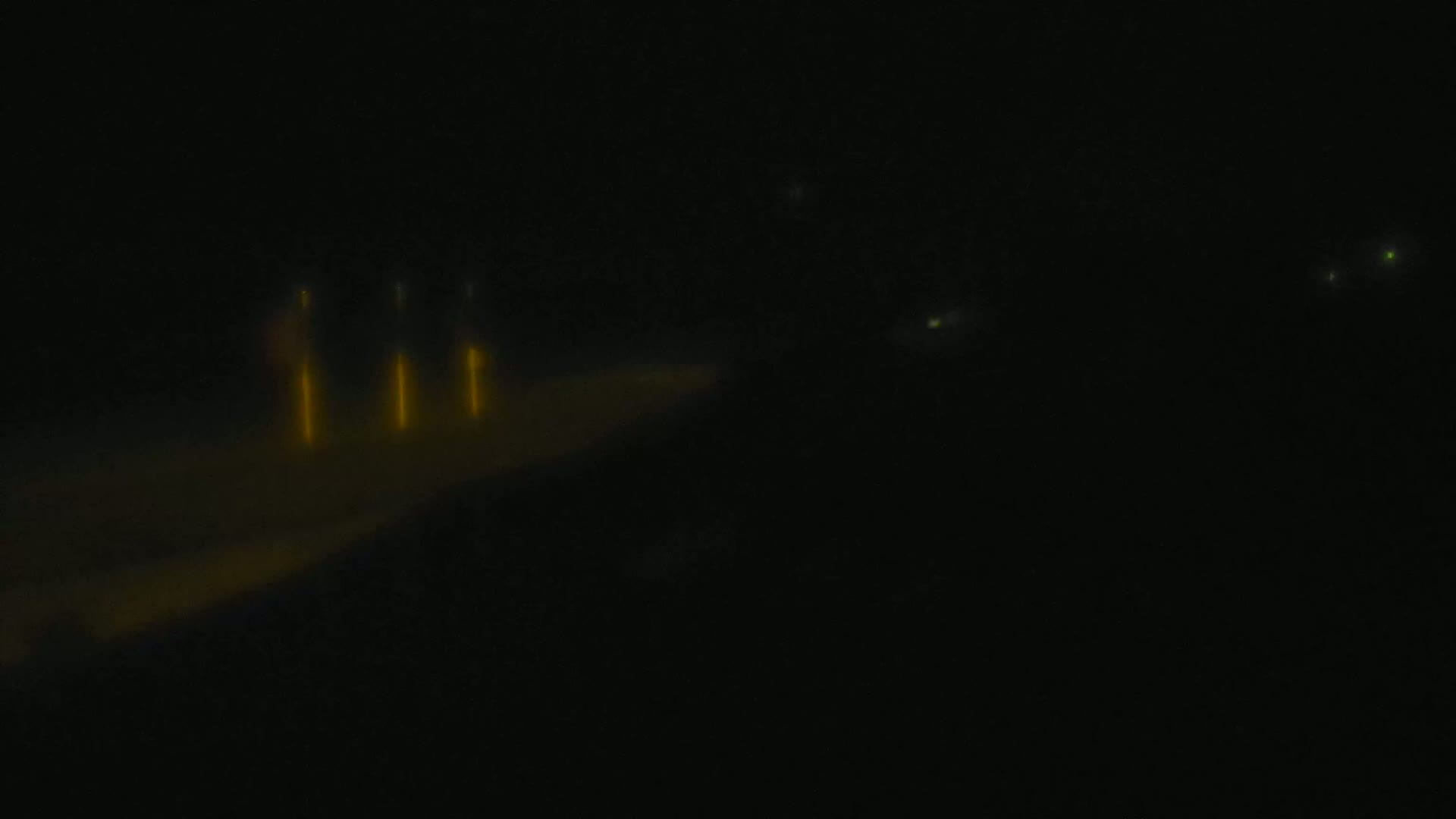 A blurry, dark image shows a few distant, bright yellow lights, possibly from vehicles, on a dark road or runway at night.