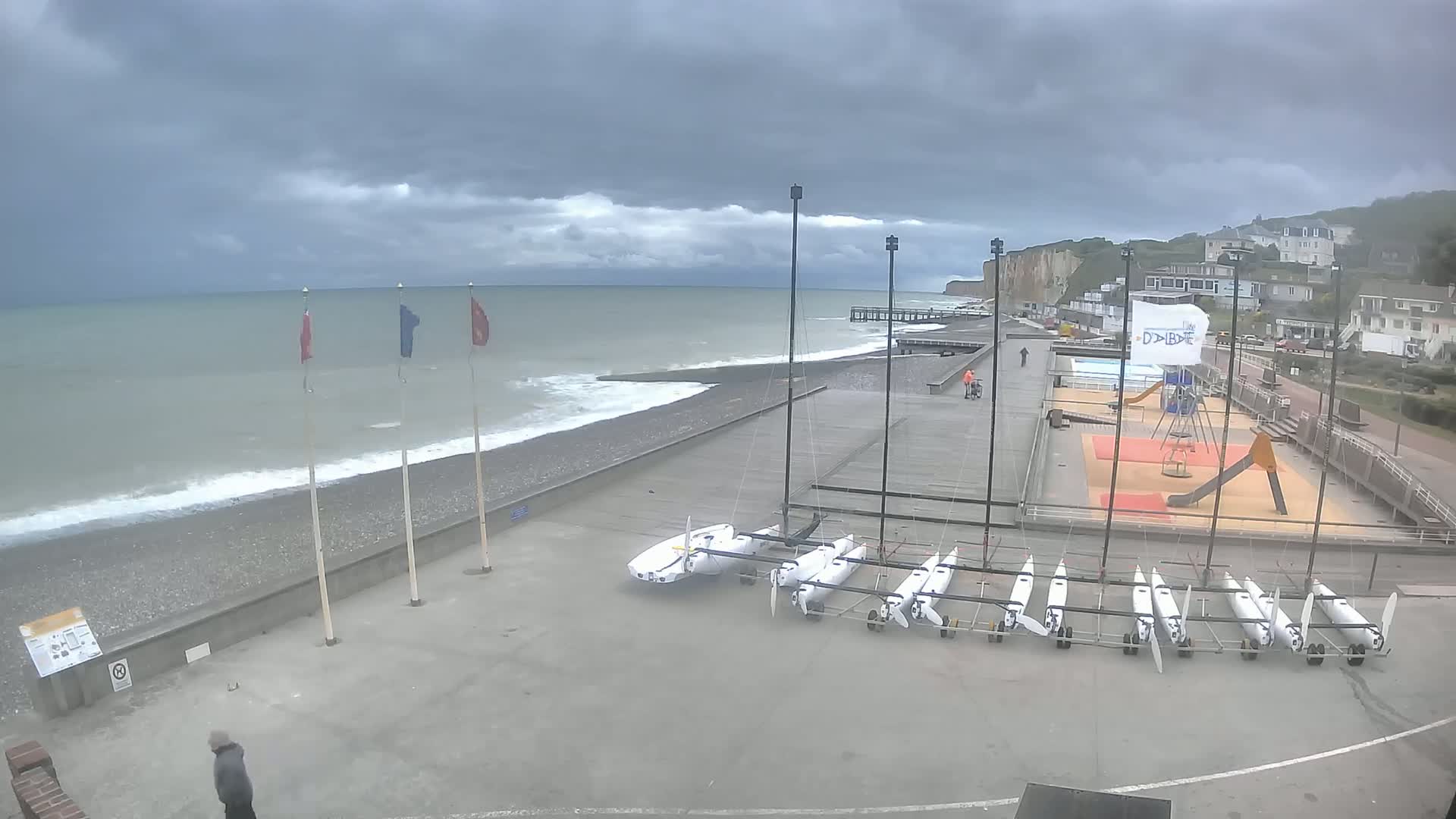 Several small sailboats are stored on a paved area next to a rocky beach with a gray, cloudy sky and a choppy sea.
