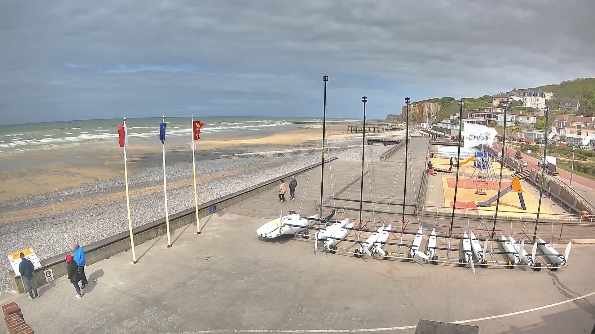 A windy day at a beach with several small sailboats parked near a playground and a few people walking along a paved path next to the shore.