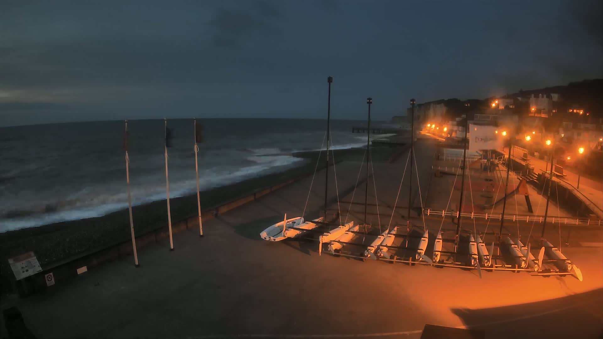 Several small sailboats are stored on a beach at night near a cliffside town under a dark, cloudy sky with somewhat choppy waves.