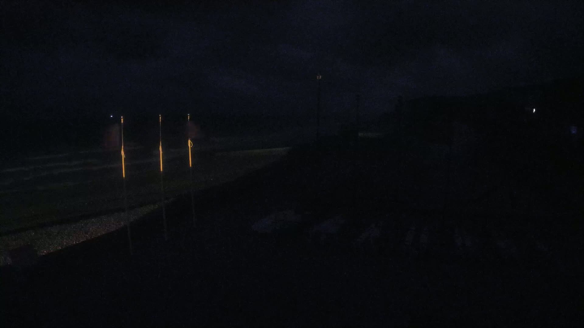 At night, several illuminated poles stand near a dark, seemingly grassy area next to a body of water under a cloudy sky.