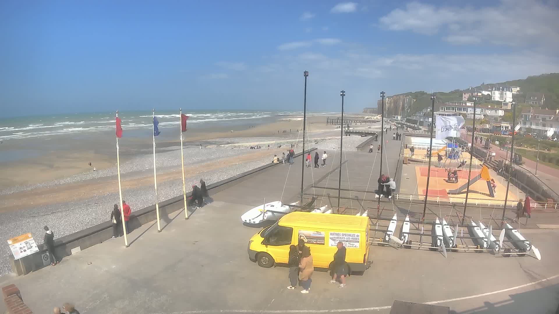 A mostly sunny day at a beach with a pebble shore, showing people walking along a paved area with several small sailboats and a yellow van parked nearby, and white-cliffed buildings in the background.