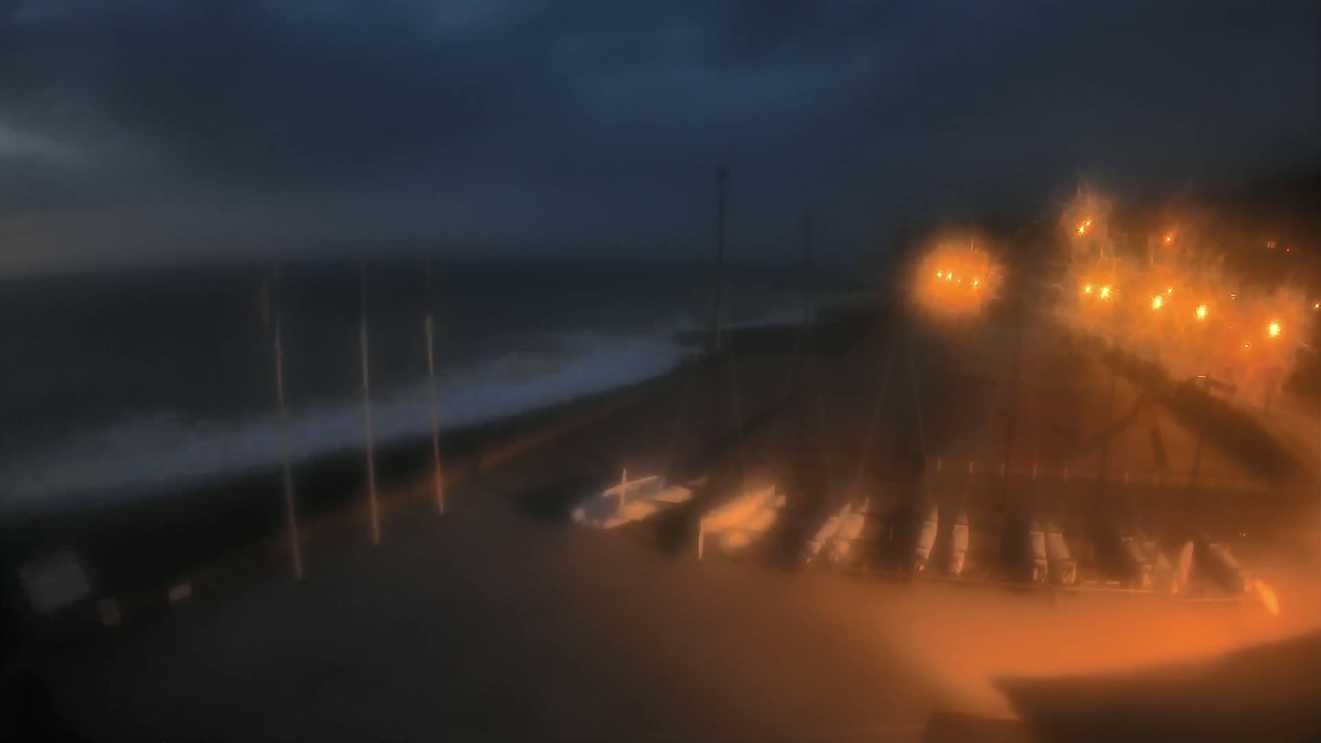A nighttime, rain-blurred view shows a dark ocean with waves breaking on a shore where several small boats are parked near lit structures.