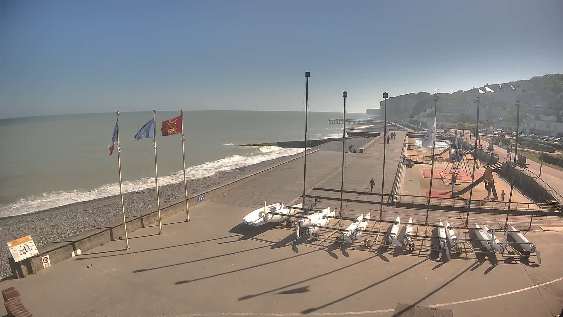 A sunny day at a seaside location shows a paved area with small sailboats, a playground, and a pier extending into a wavy ocean under a clear sky.