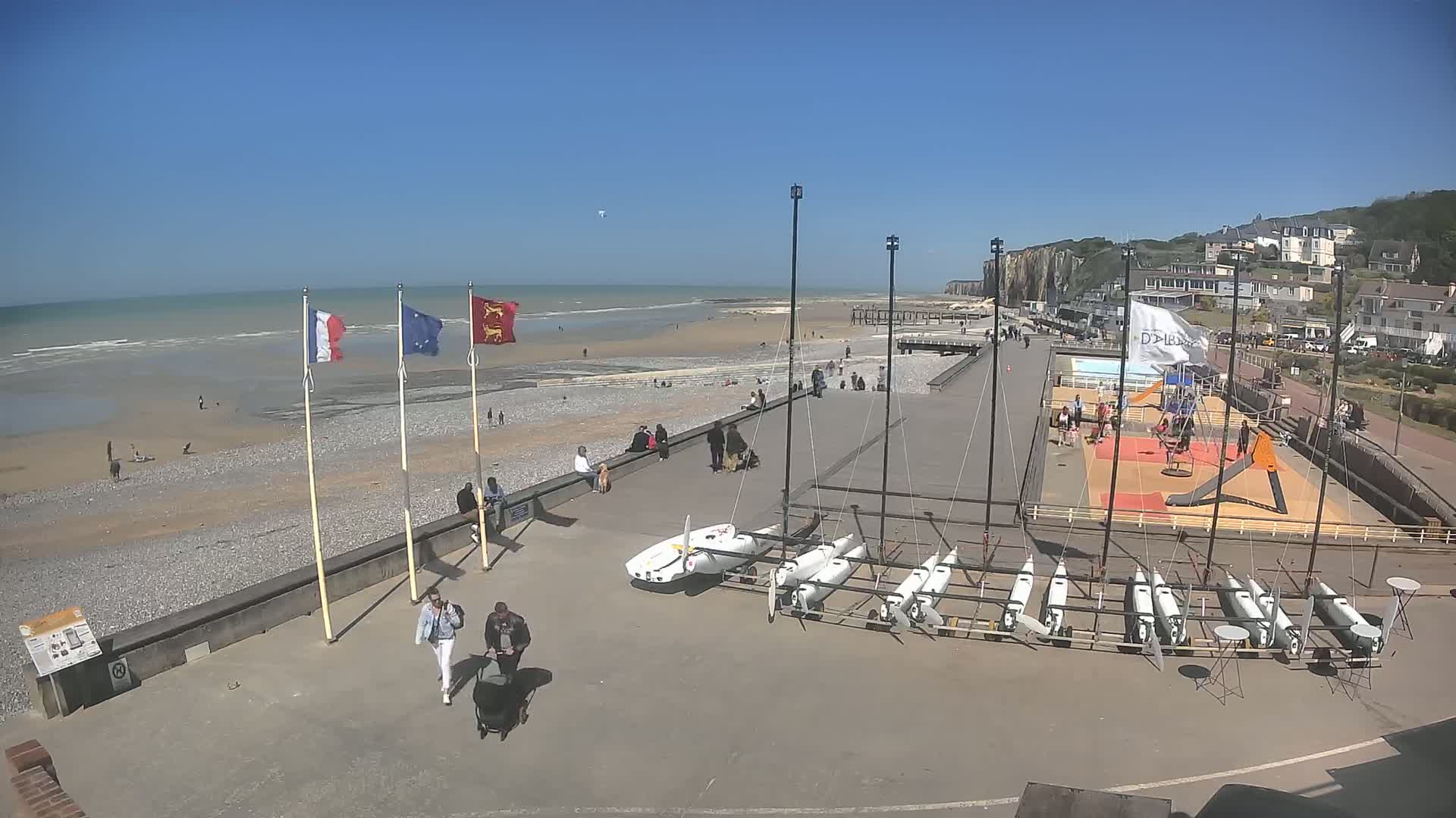 A sunny beach scene shows a pebbled shore, a boardwalk with people, several small sailboats on land, and a playground, all situated next to white cliffs.