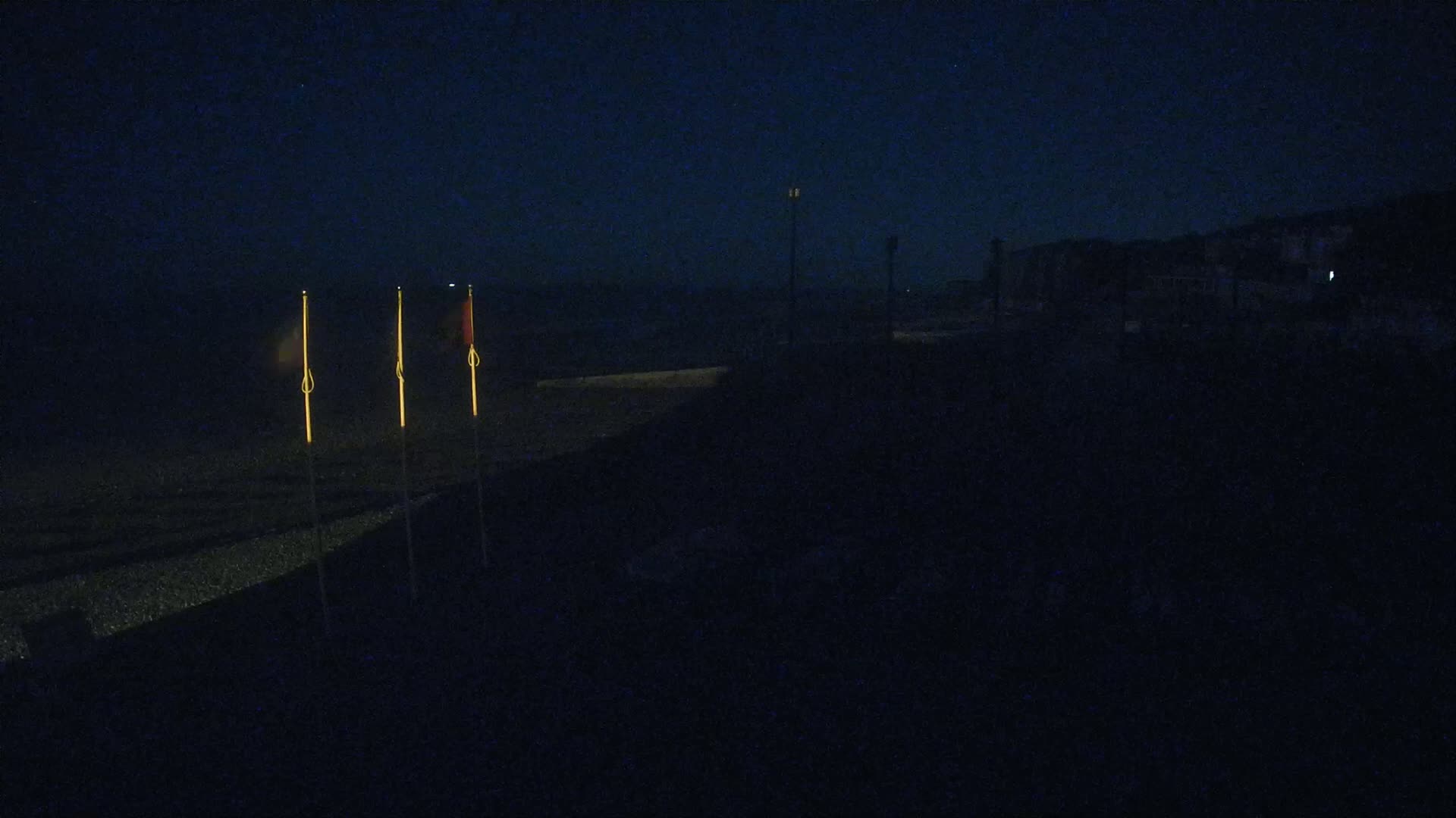 Three flagpoles with dimly lit flags stand on a dark beach at night.