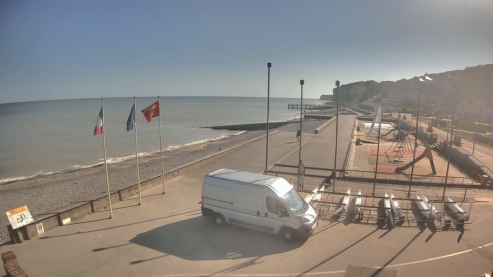 A sunny day at a seaside location shows a van parked near several small boats, a playground, and a shoreline with flags and a pier extending into a calm sea with whitecaps near the shore.