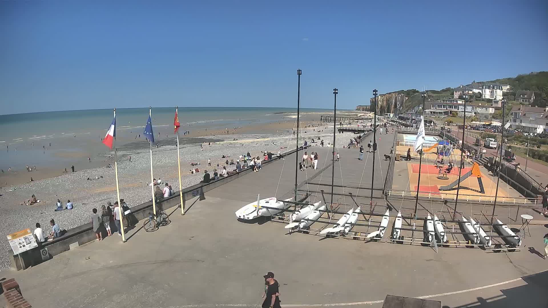 A sunny beach scene shows many people relaxing and swimming in the ocean, with several sailboats stored on the shore near a playground and a promenade.