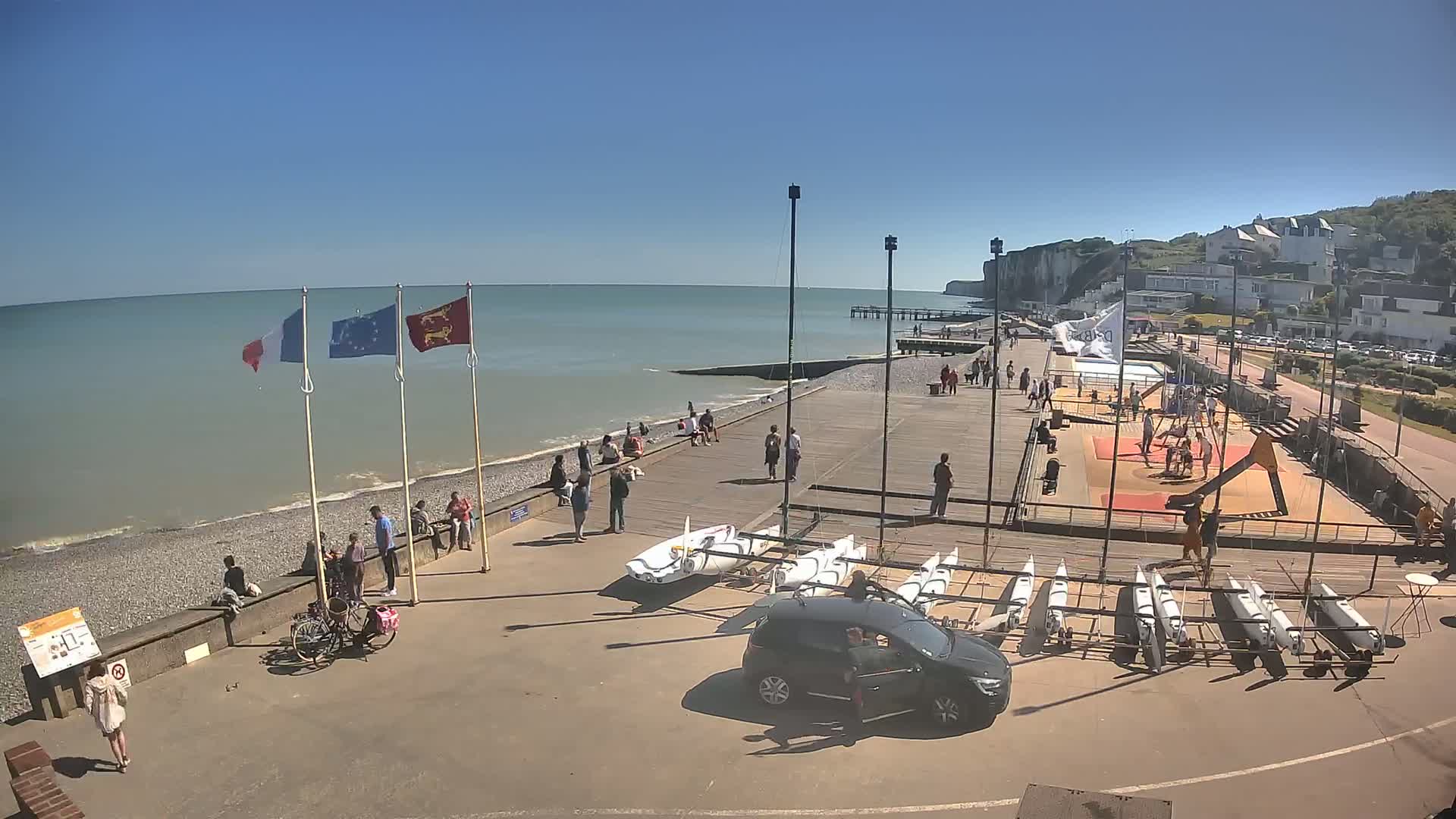 A sunny day at a seaside location shows people relaxing and strolling along a boardwalk near the ocean, with sailboats stored nearby and white cliffs visible in the background.
