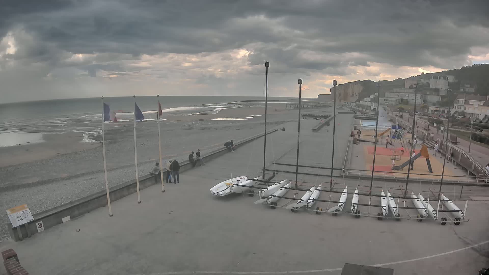 A mostly cloudy sky hangs over a beach with several people, sailboats, and a playground; the ocean is calm and receding, exposing a wide expanse of sand and shallow water.