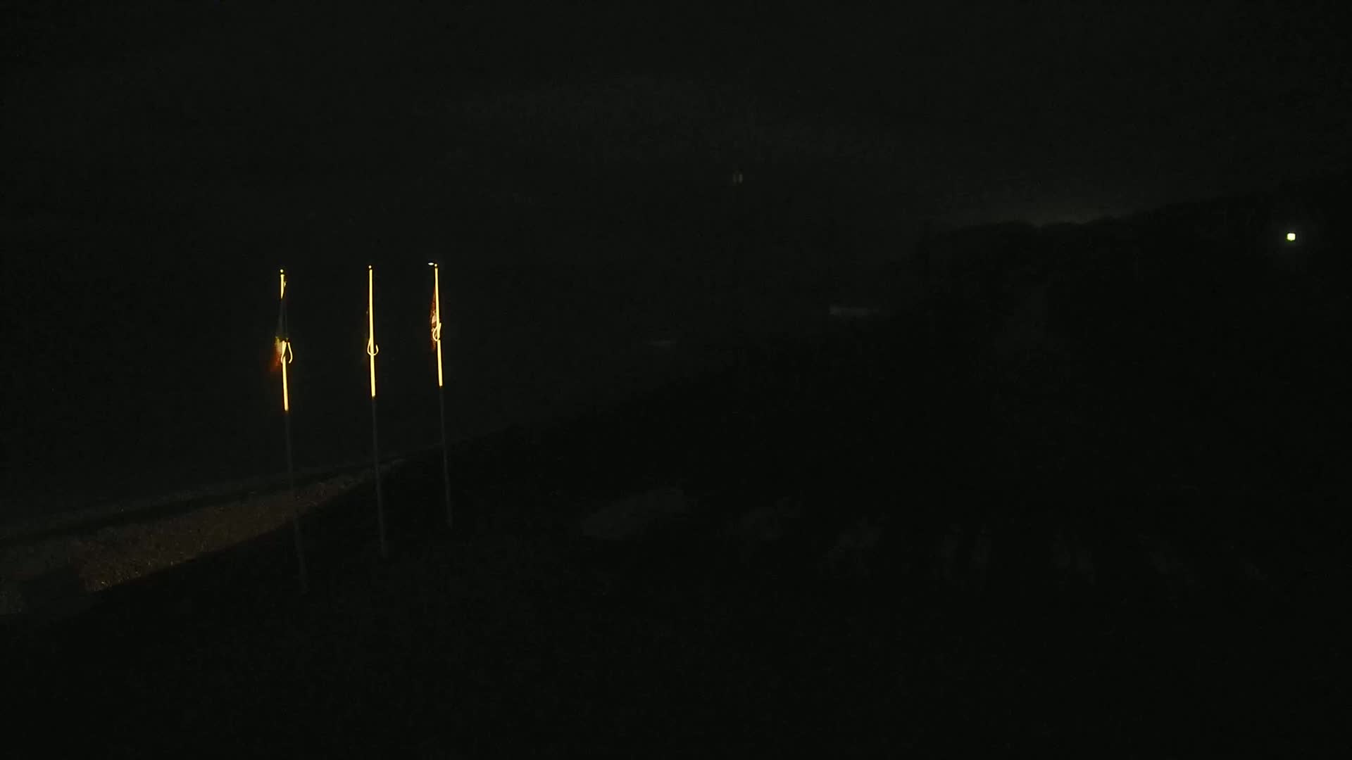 Three lit, thin, vertical poles stand against a dark, nighttime background.