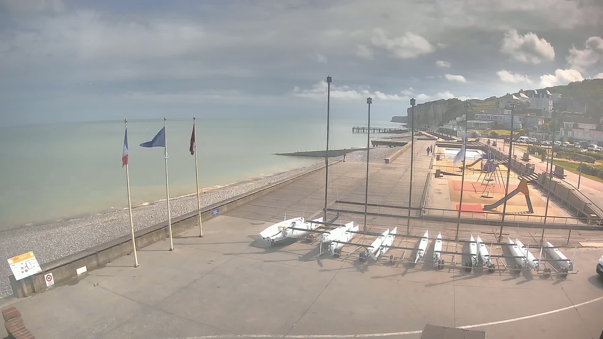 A seaside area with a calm sea under a partly cloudy sky features several sailboats stored on the shore near a playground and a walkway leading to white cliffs in the distance.
