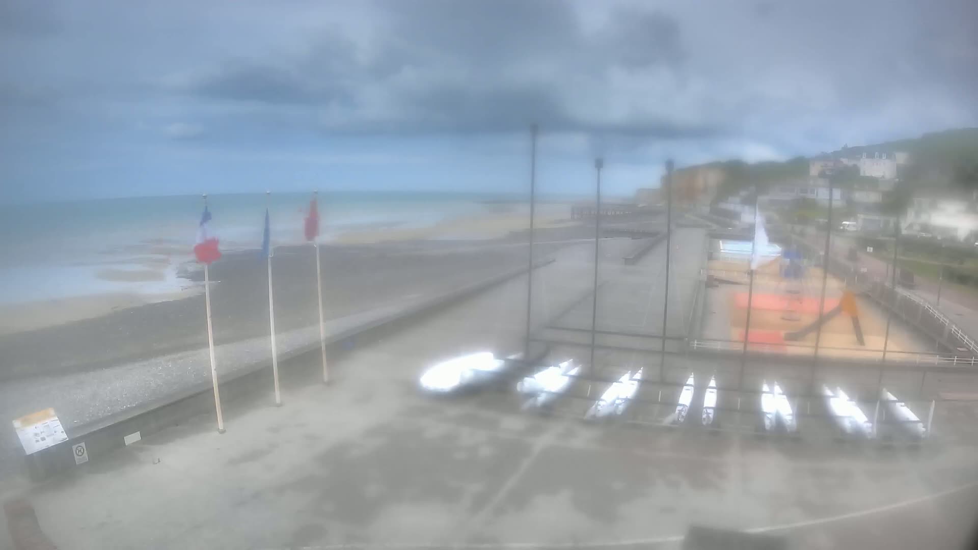 A hazy, overcast day reveals a beach with several sailboats stored near a playground, overlooking a calm sea with three flags in the foreground.