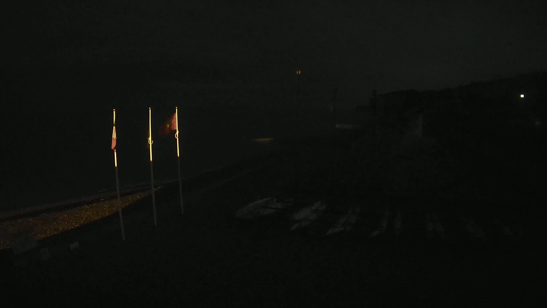 Three illuminated flags stand on a dark, nighttime shoreline, with a dimly lit structure visible in the distance.