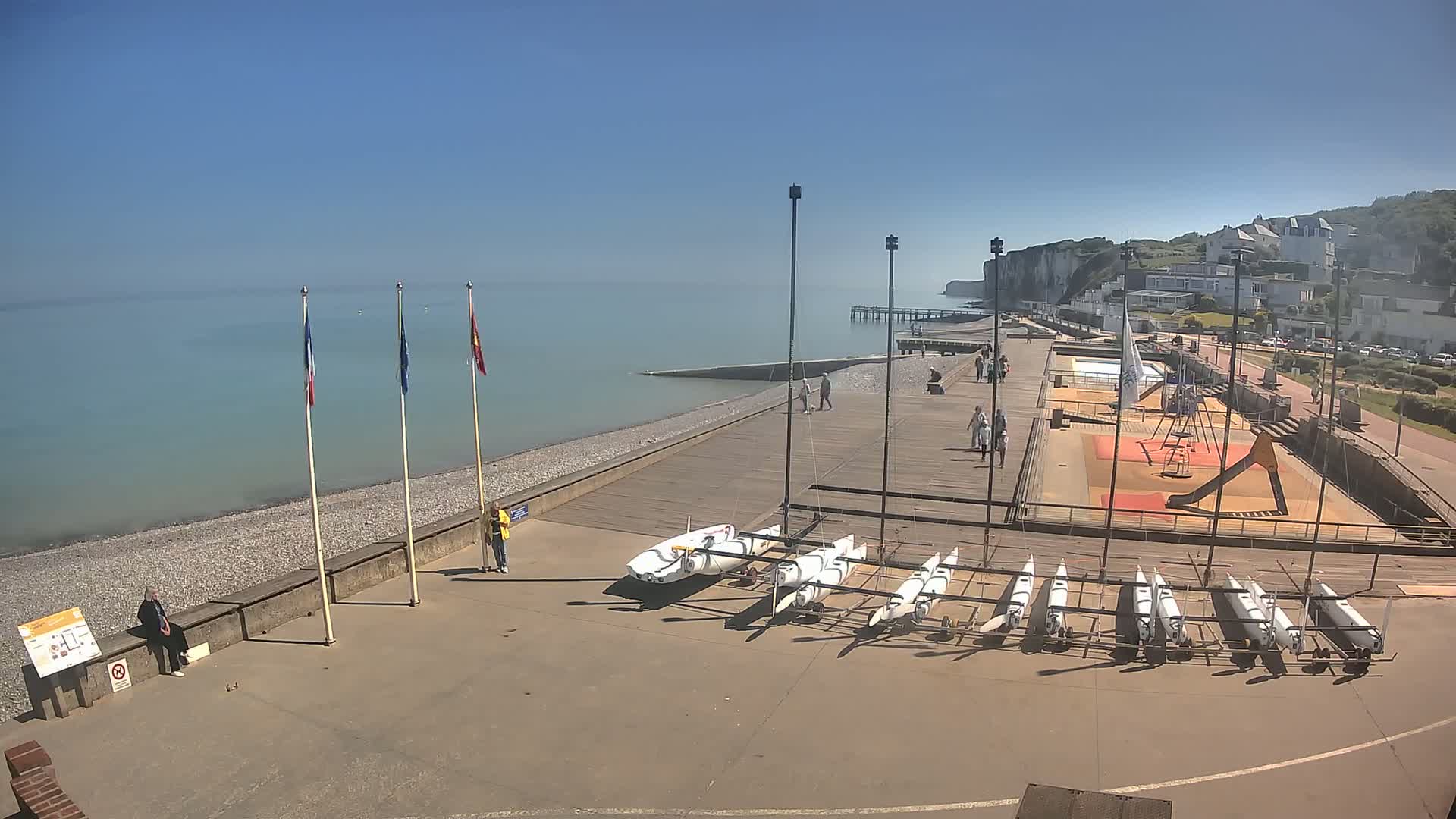 A sunny day at a seaside town shows a pebble beach, a boardwalk with several small sailboats stored, a playground, a pier extending into a calm sea, and white cliffs in the background.