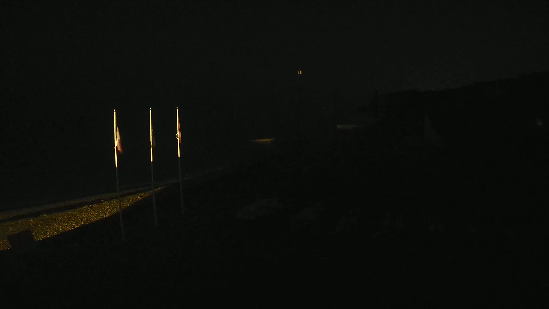 Three flags are illuminated on poles at night, standing before a dark, seemingly beach-like area.