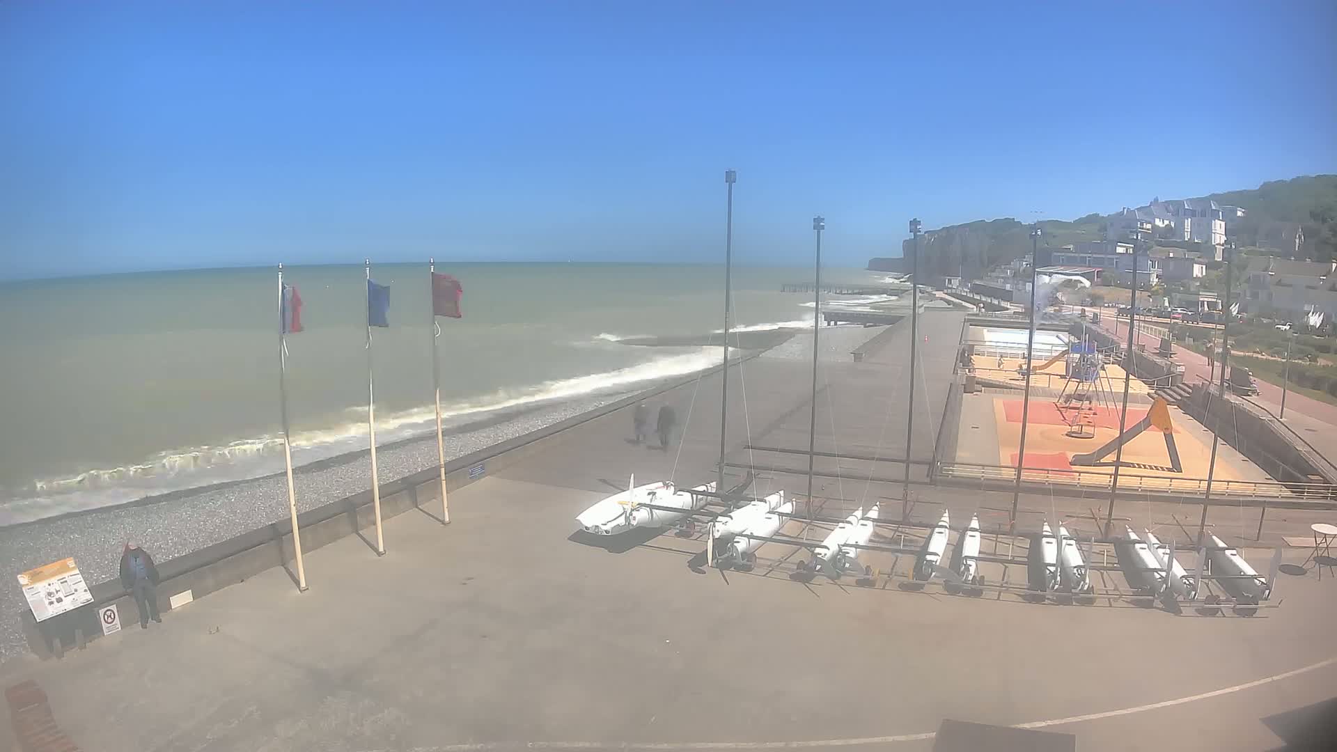 A beachside area with several small sailboats stored on land, a playground, and a few people walking along a pebble shore under a clear, sunny sky with some small waves breaking on the beach.